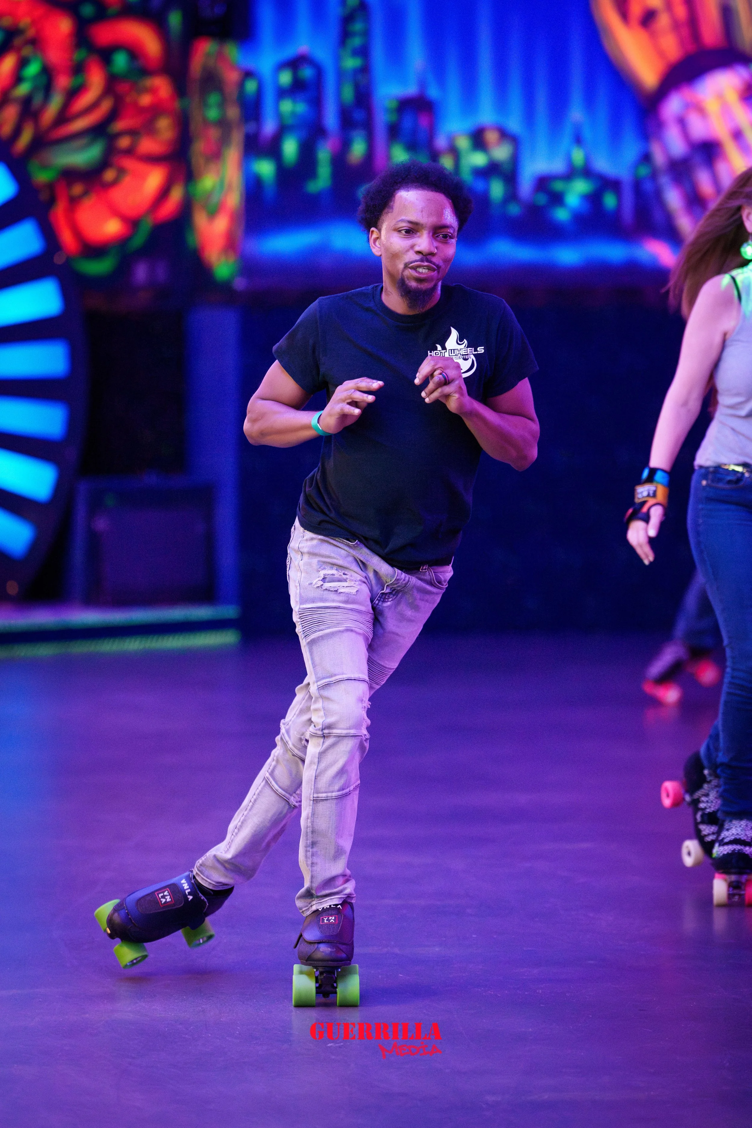 A man roller skating indoors with vibrant, colorful mural artwork in the background. He is wearing a black t-shirt and light jeans, and appears to be enjoying himself.