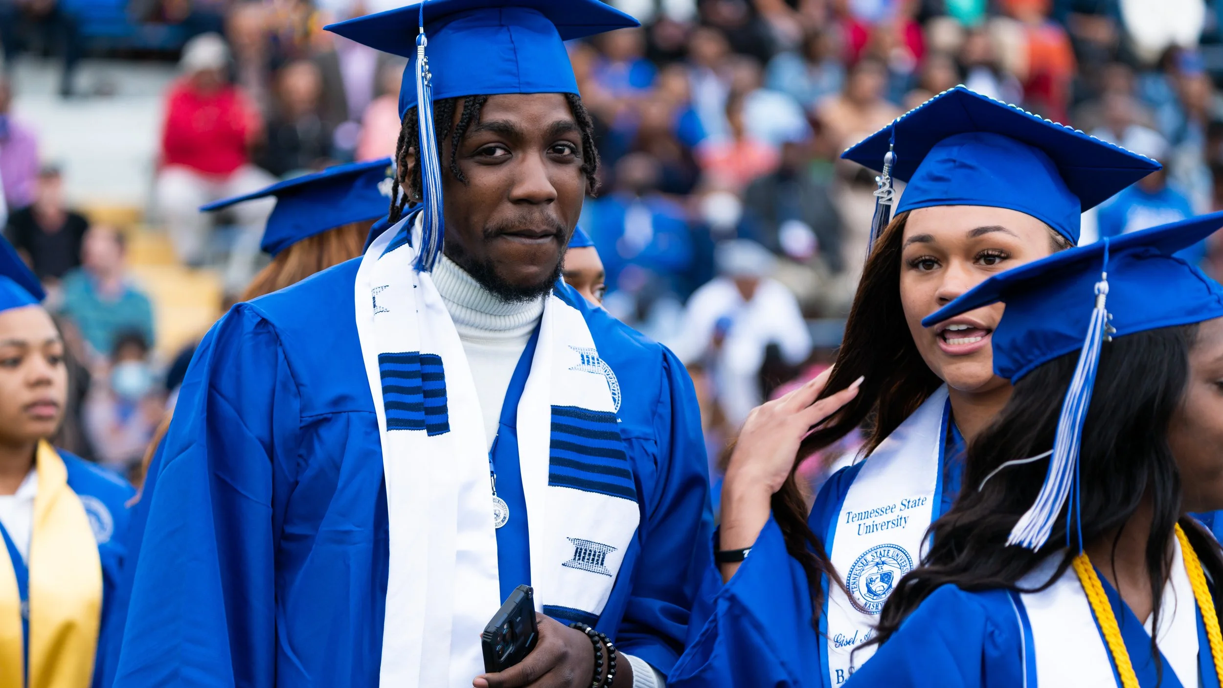 Graduates in blue caps and gowns at a university commencement ceremony, with a crowd in the background.