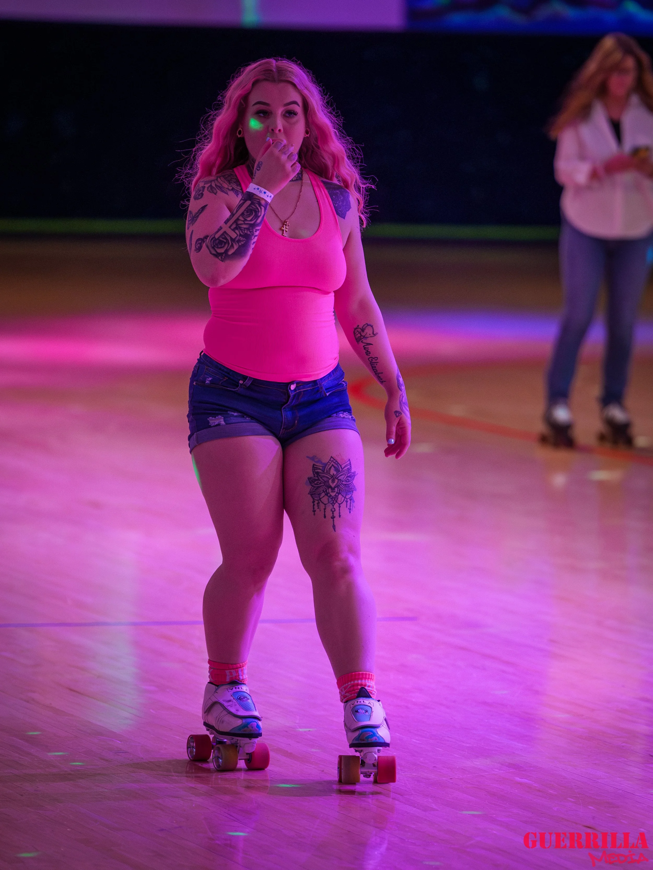 A woman roller skating on a wooden rink illuminated by pink and purple lights. She is wearing a pink tank top, denim shorts, and sneakers. She has tattoos on her arms and legs, and is covering her mouth with her hand while looking at the camera. In t
