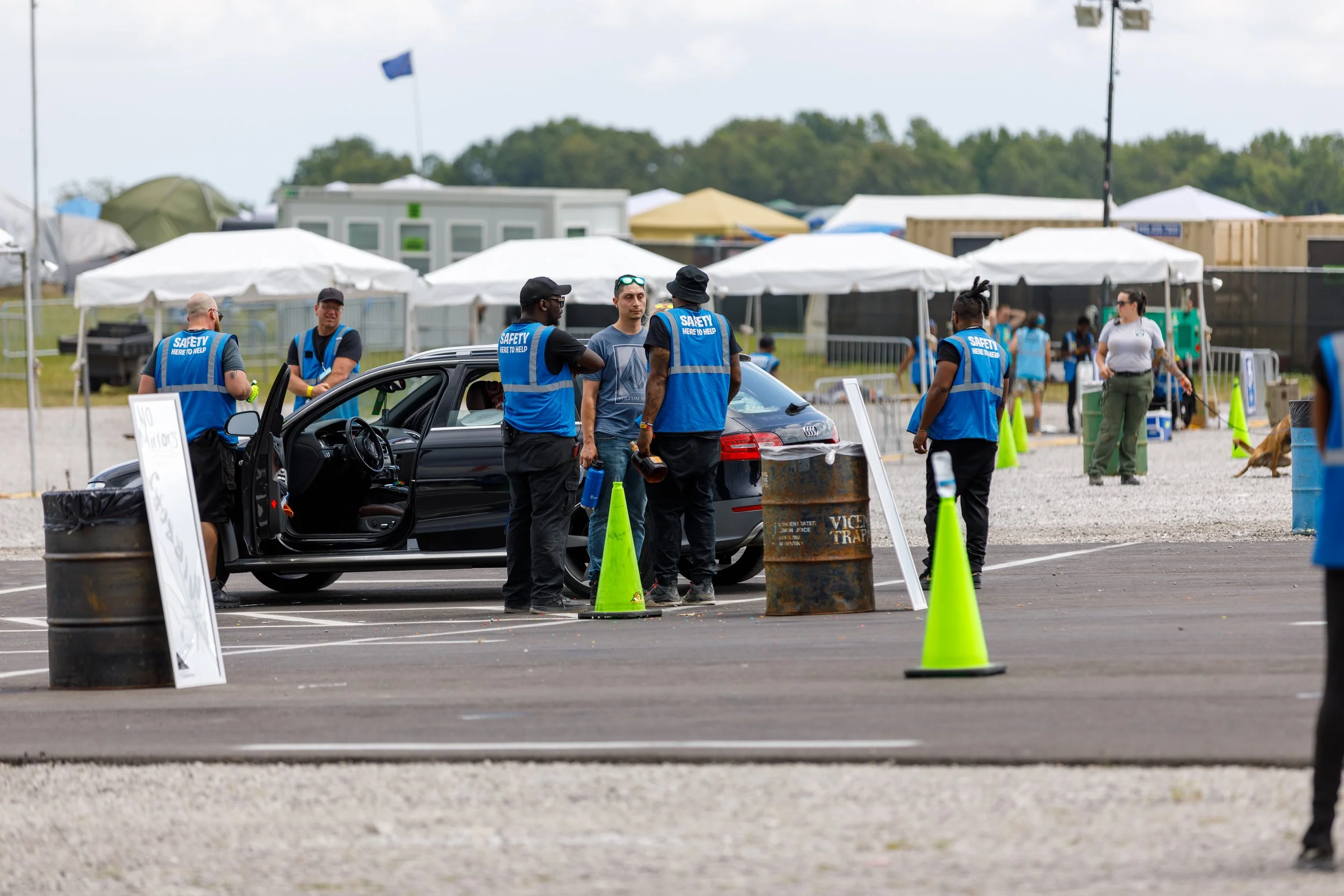 Security personnel in blue vests talking to a man at a car during an outdoor event, with tents and people in the background.