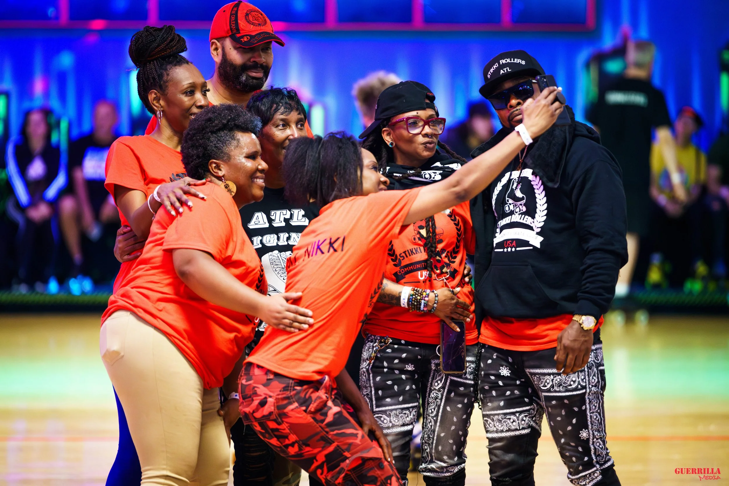 A group of people, mostly women, taking a selfie together in a gymnasium. They are wearing matching orange shirts with some text, and some are wearing black and orange outfits. The background features colorful lighting and other people in the gym.