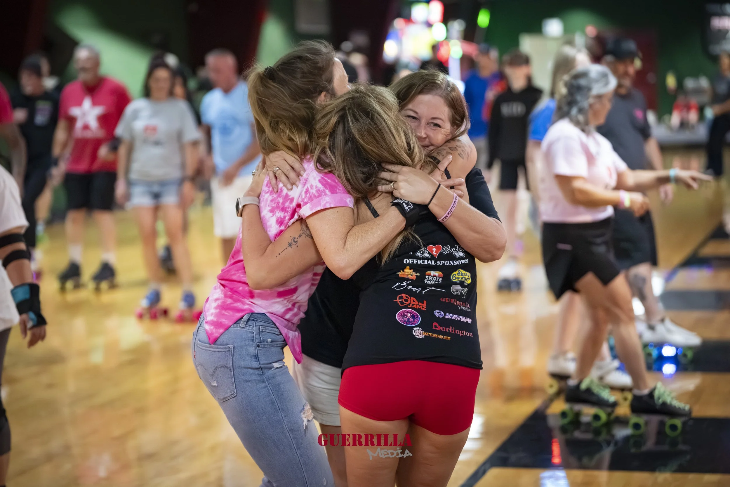 Three women hugging and celebrating at a roller skating rink. One woman is wearing a black shirt with logos, another in pink tie-dye shirt, and the third in a black T-shirt and red shorts. Other skaters are visible in the background.