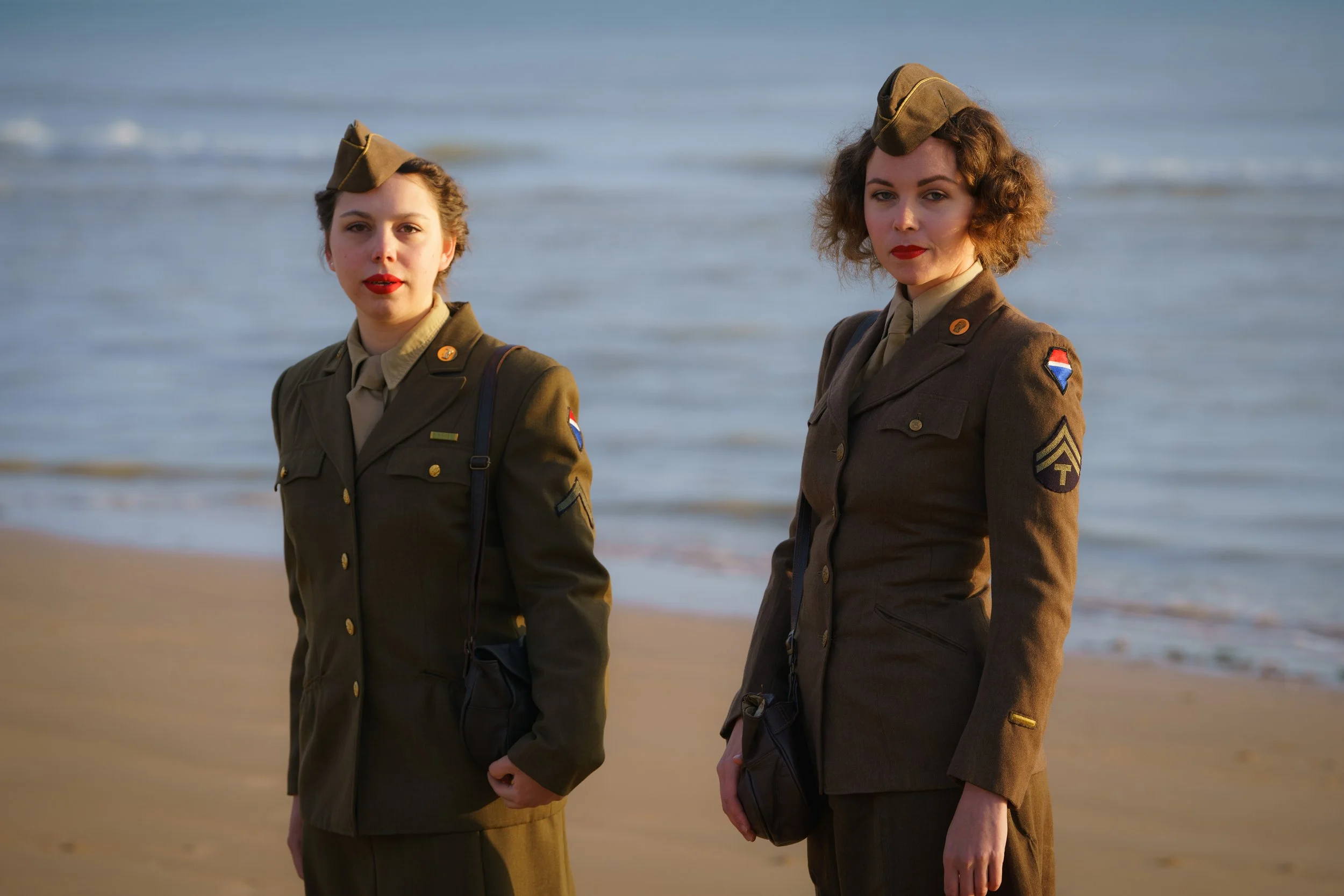 Two women dressed in military uniforms standing on the beach with the ocean in the background.