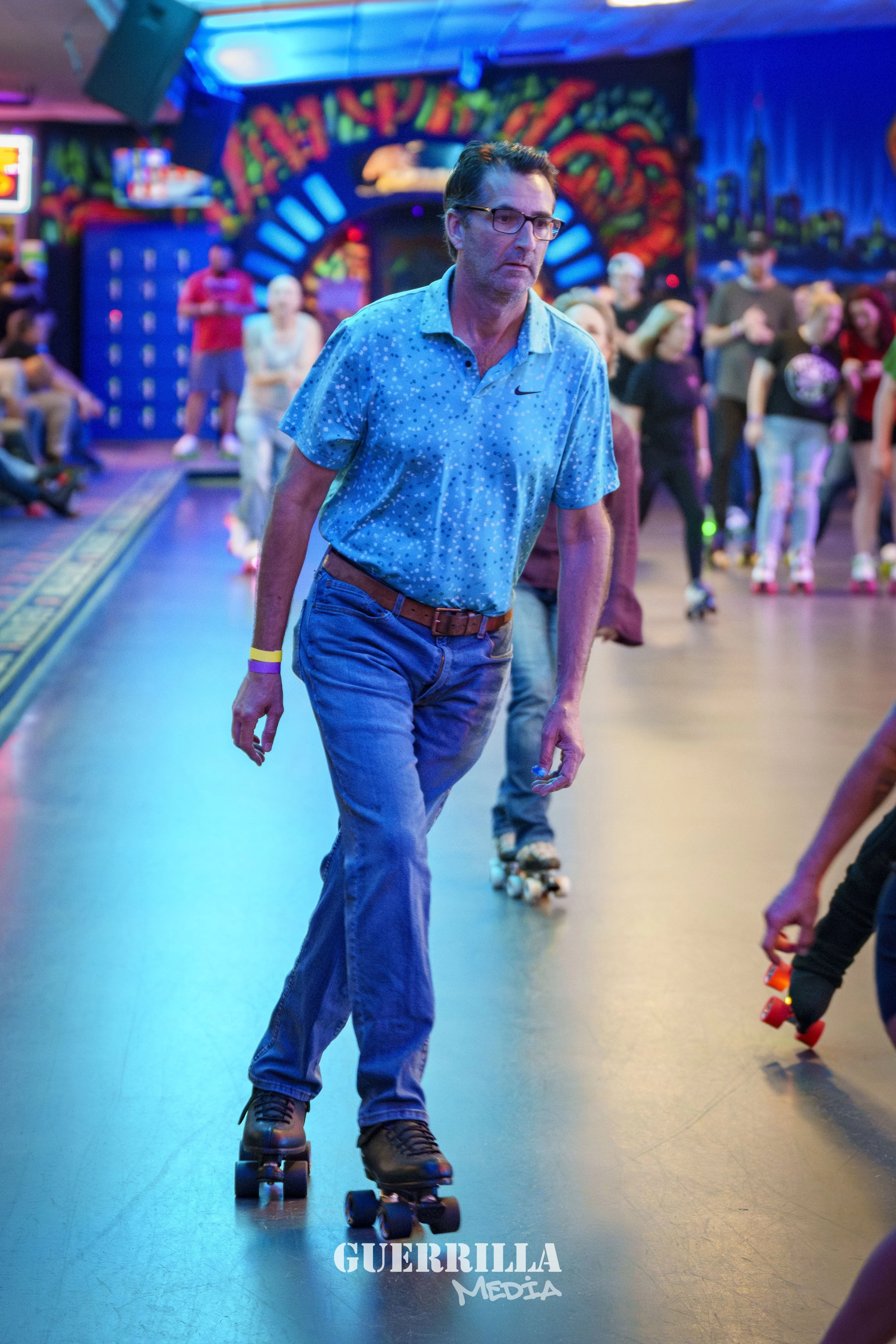 A man roller skating inside an arcade or roller rink with colorful lights and graffiti-style wall art in the background. Other skaters and spectators are visible.