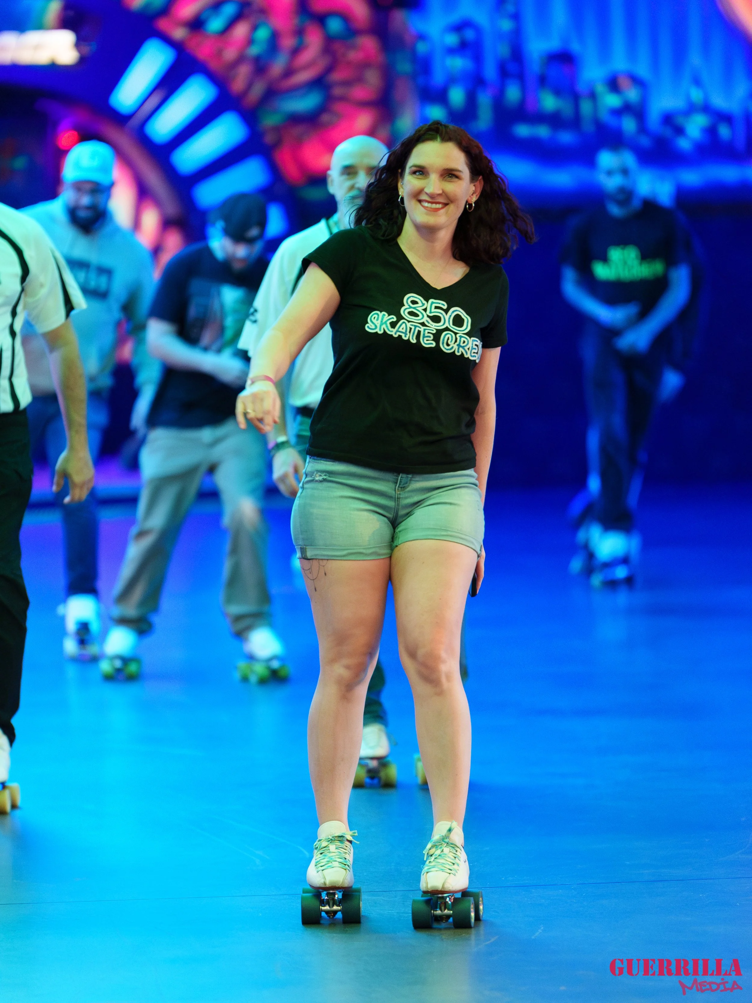 Woman roller skating indoors at a rink with colorful background, smiling, wearing a black T-shirt.