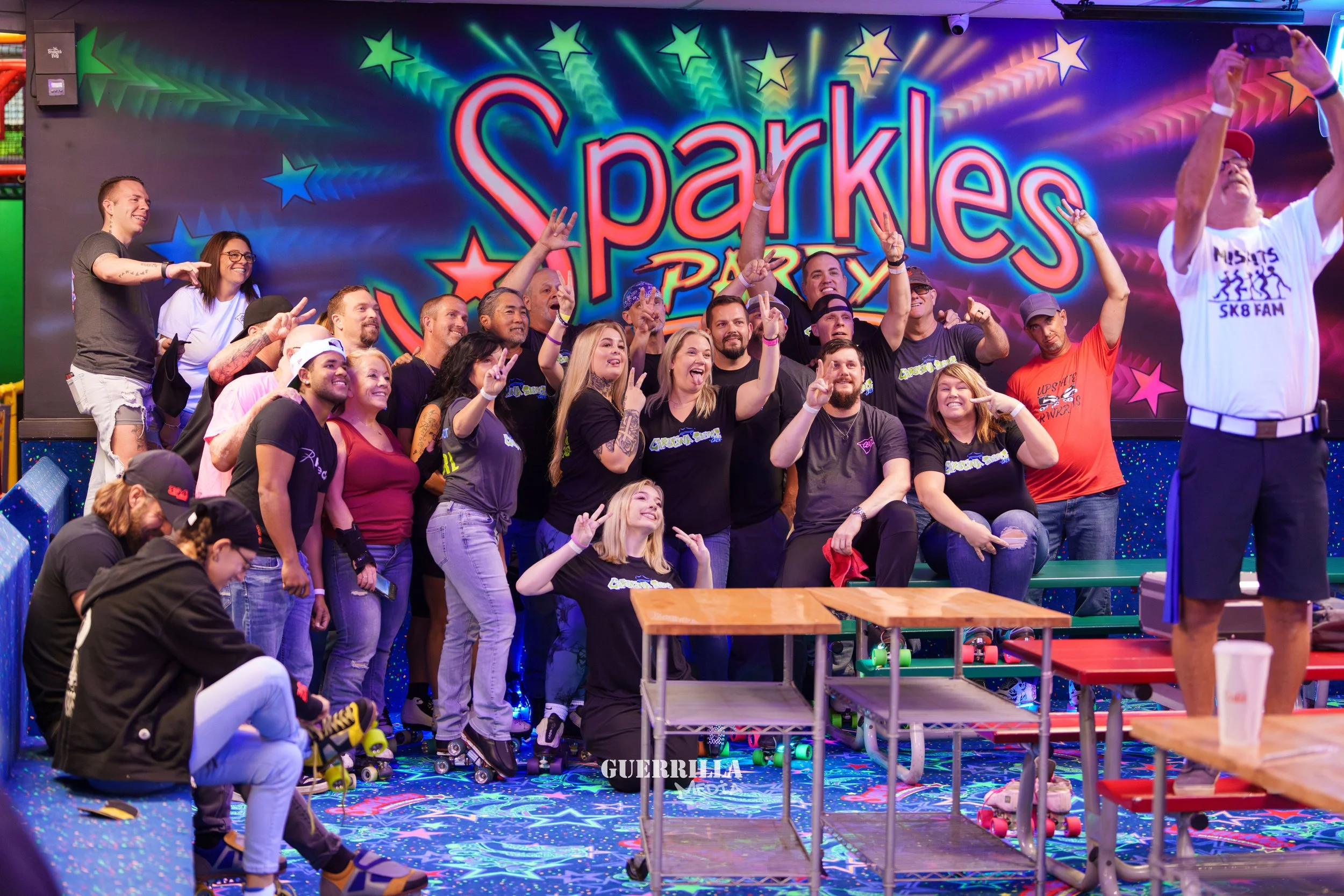 A group of people posing and smiling in front of a colorful sign that reads 'Sparkles Party' at an indoor roller skating rink. Many are wearing black shirts with a logo, and some are on their skates. The background features a vibrant, decorated wall 