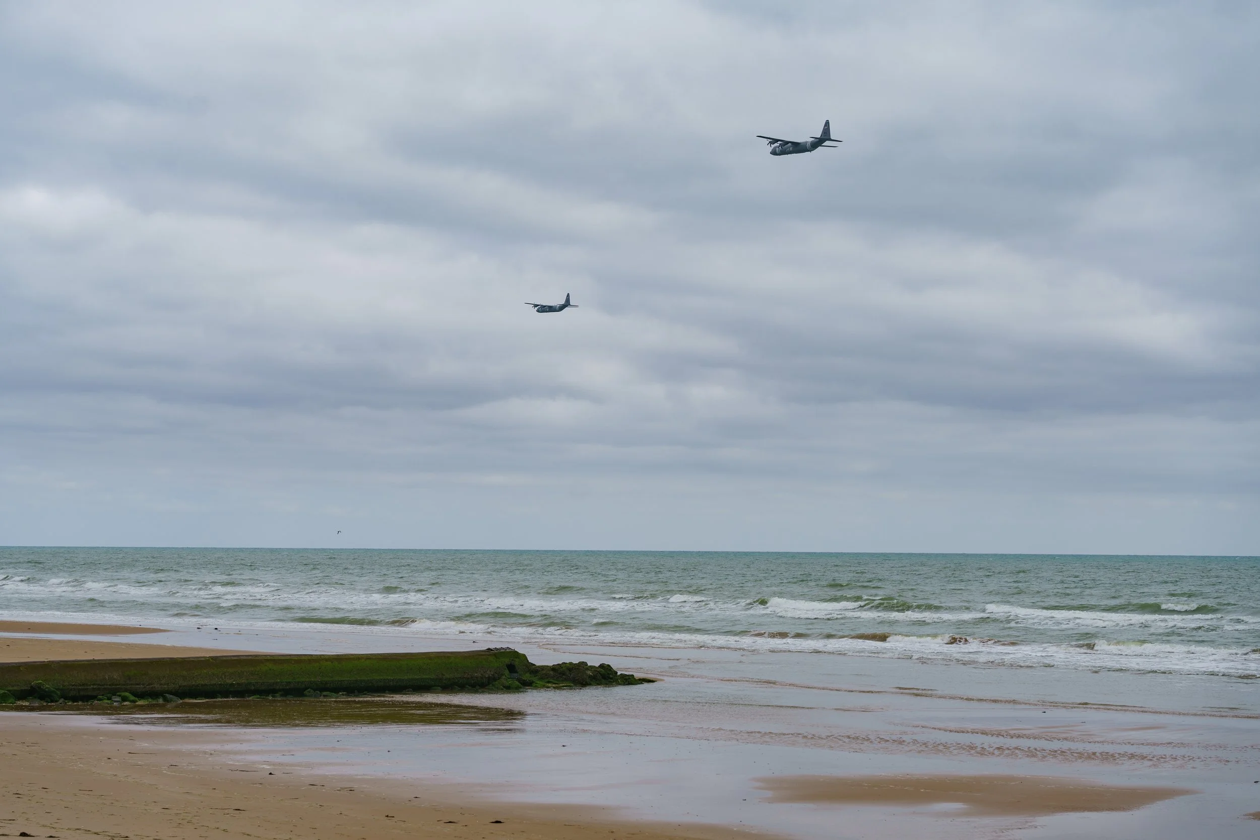 Overcast cloudy sky with two airplanes flying over the ocean near a sandy beach with some rocks and moss.