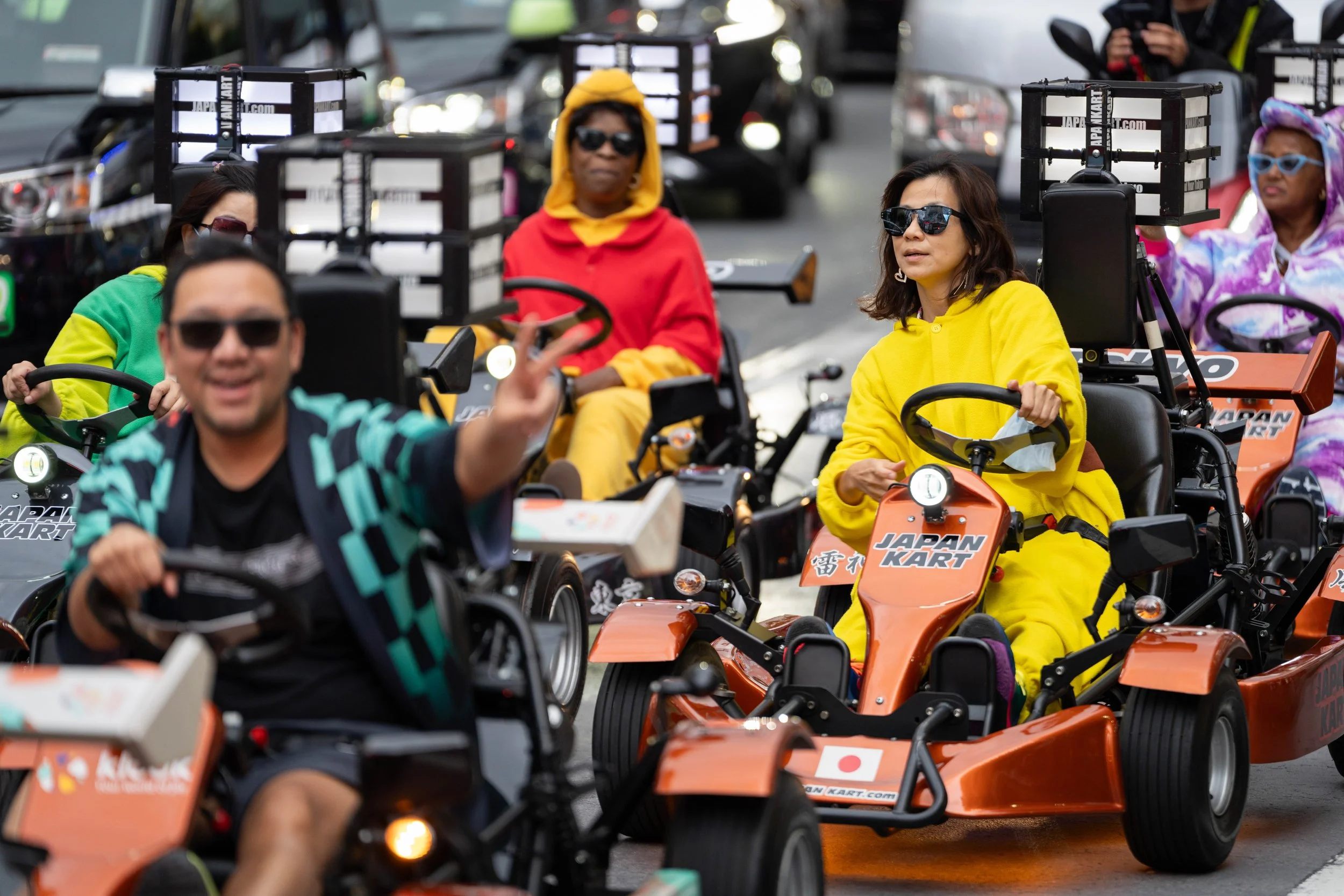 Group of people riding orange go-karts on a city street, wearing colorful clothing and sunglasses, some waving and smiling.