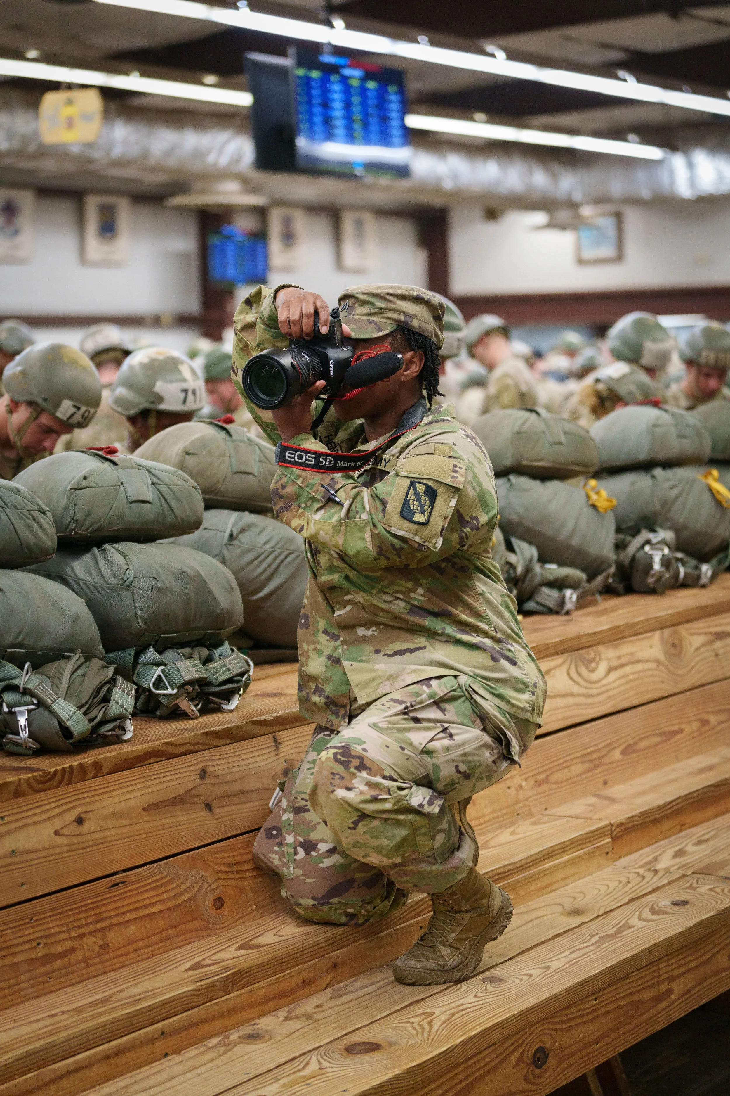 A soldier in camouflage uniform kneeling on a wooden bench, taking a photograph with a professional camera inside a room filled with other soldiers in uniform and gear.
