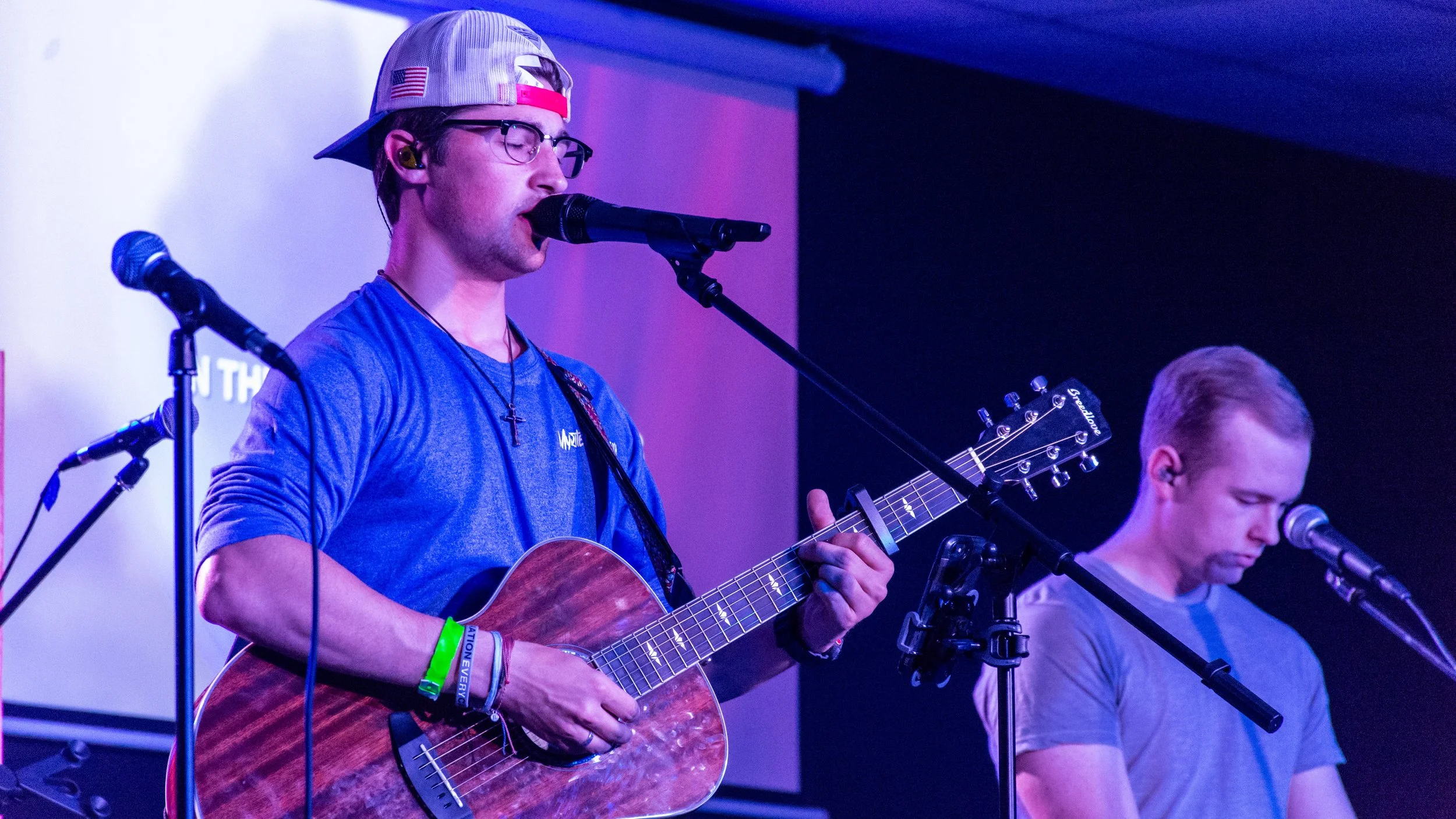 Two young men performing music on stage, one singing into a microphone and playing an acoustic guitar, the other standing with a microphone, with purple and blue stage lighting.