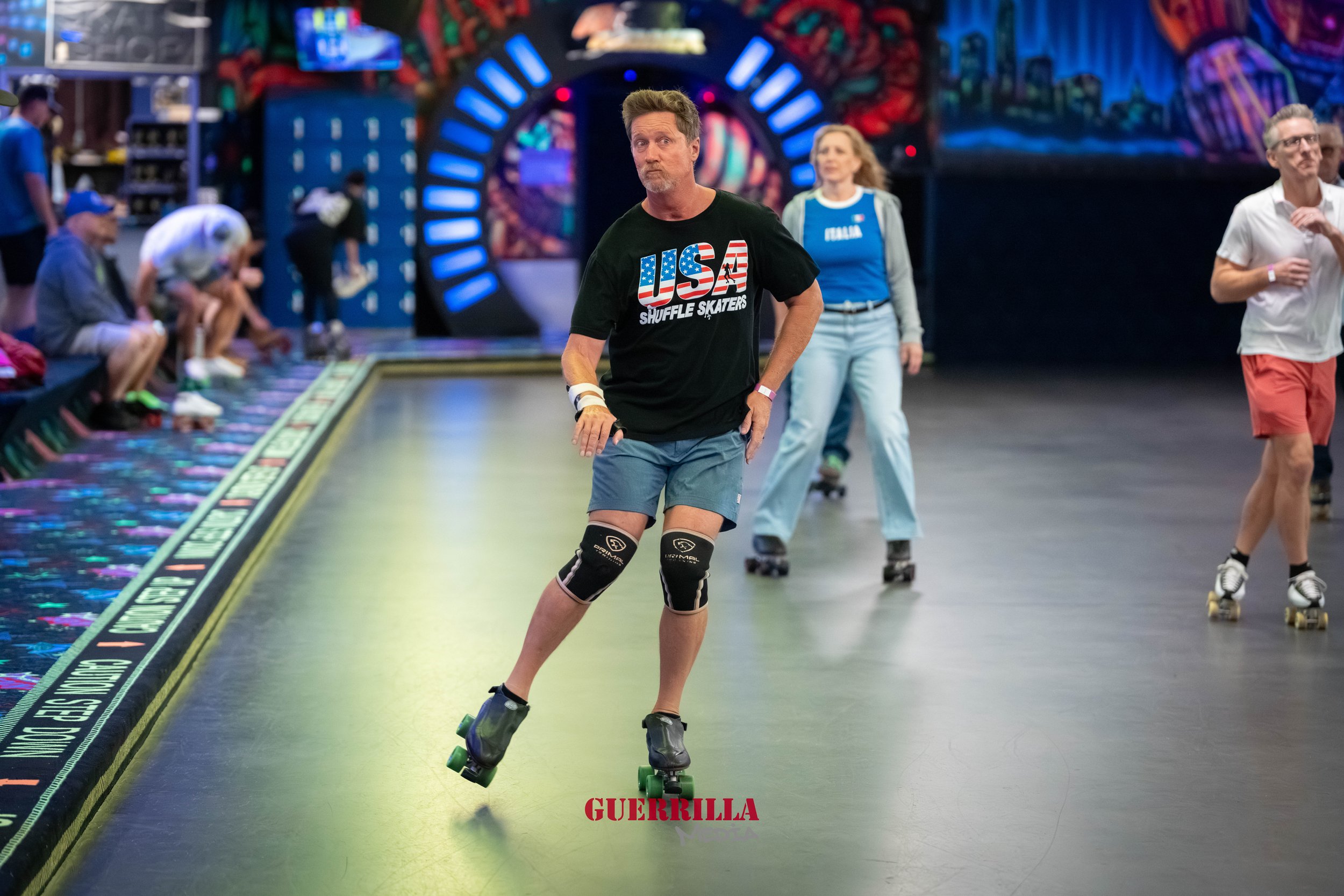 A man roller skating in an indoor skate rink, wearing a black USA shuffle skaters t-shirt, blue shorts, and knee pads. There are other skaters and spectators around him.