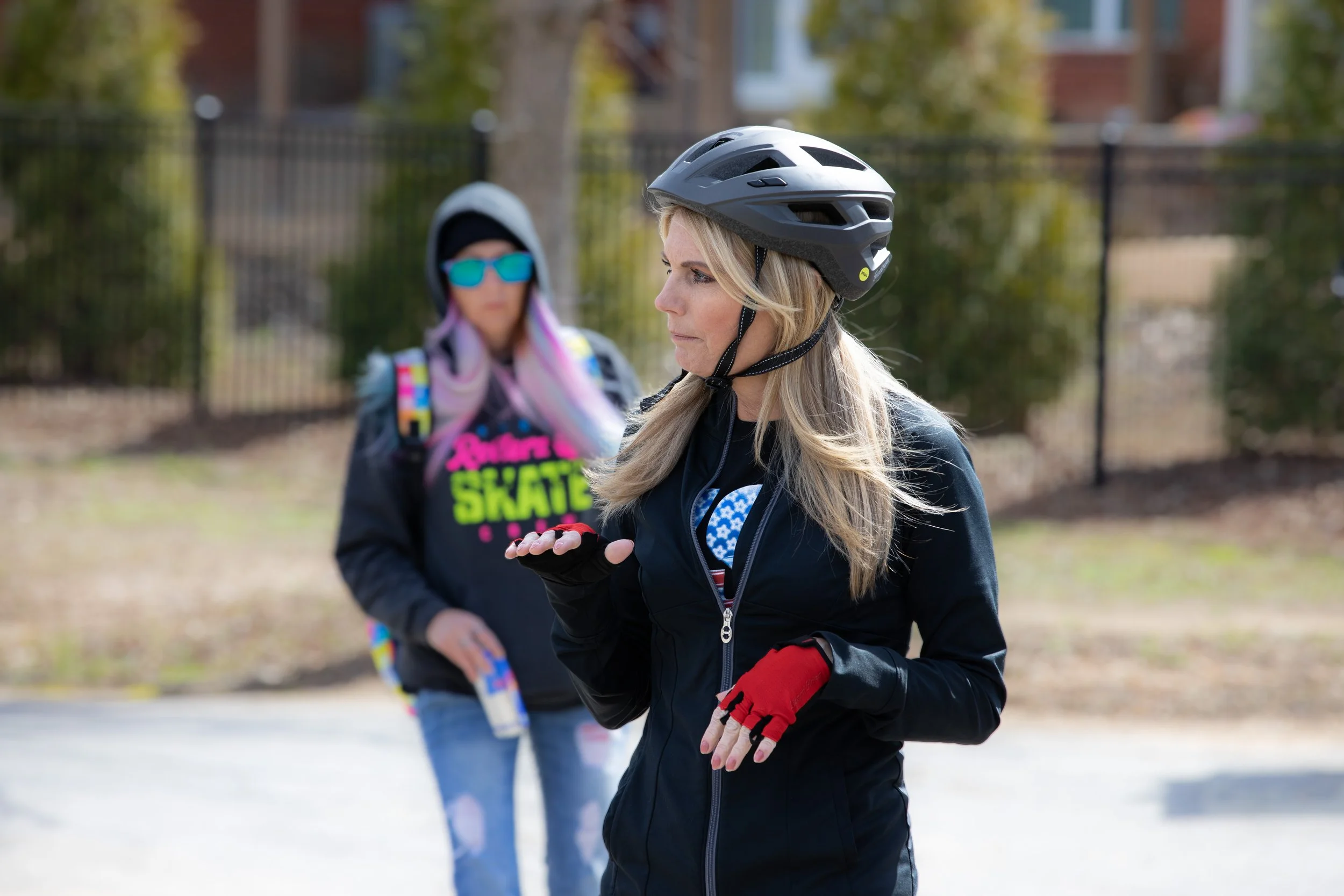 Two women wearing helmets and jackets, standing outdoors, one talking and the other listening, with trees and a fence in the background.