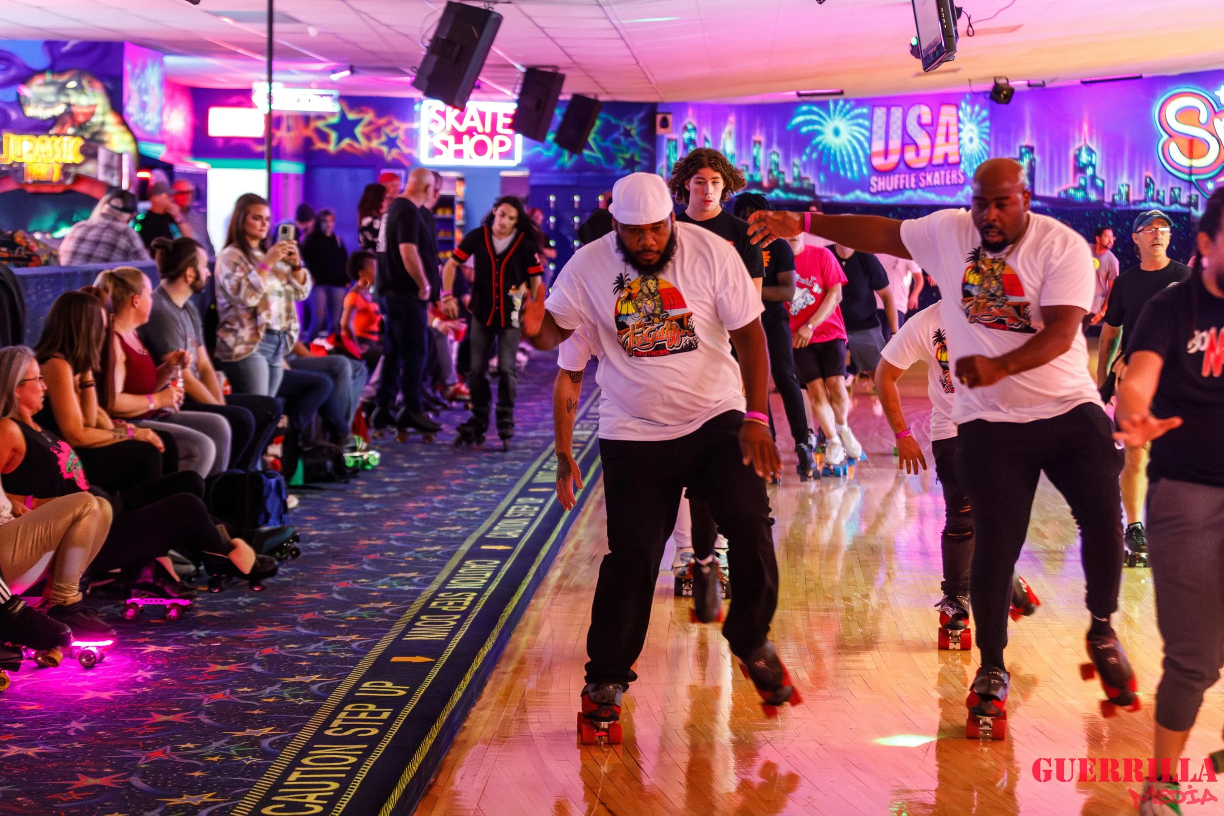 People roller skating indoors with colorful neon signs and murals, including 'Skate Shop,' 'USA Shuffle Skaters,' and 'Jurassic Park.' A row of seated spectators watches the skaters.