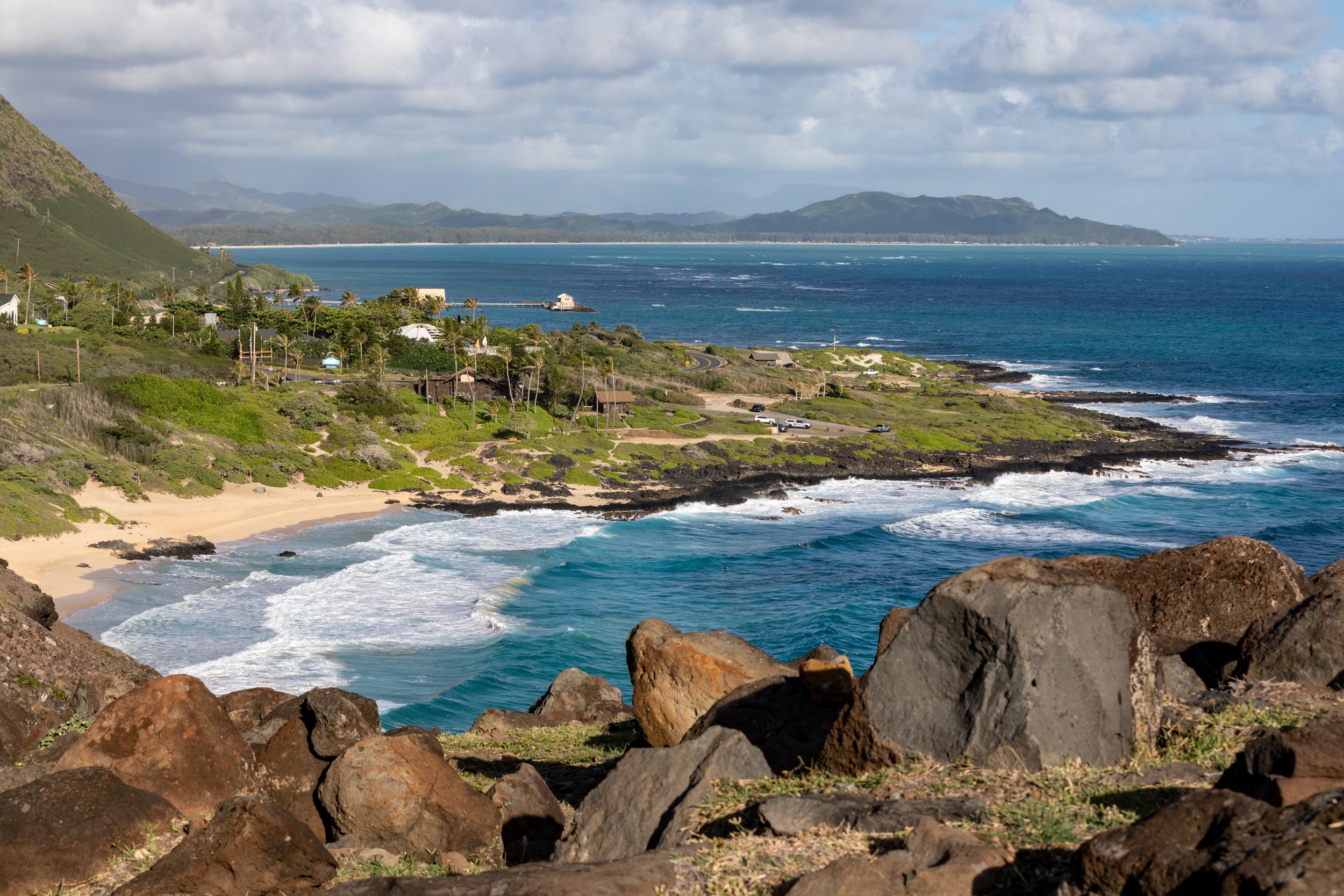 Coastal landscape with rocky foreground, sandy beach, blue ocean waves, lush green vegetation, houses, and distant mountains under partly cloudy sky in Hawaii.