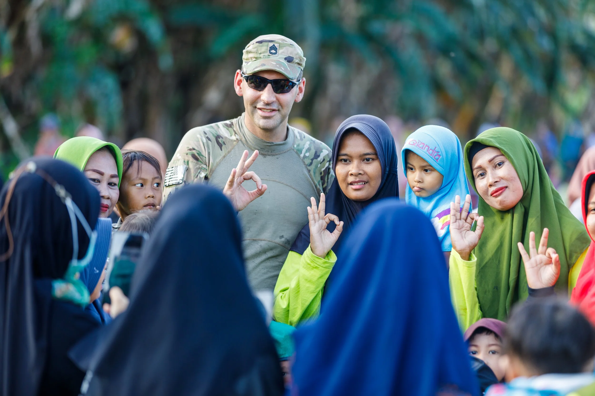 A group of diverse children and women, some wearing hijabs, gathered outdoors, listening to a man in military uniform and sunglasses, who is making an OK gesture with his hand.
