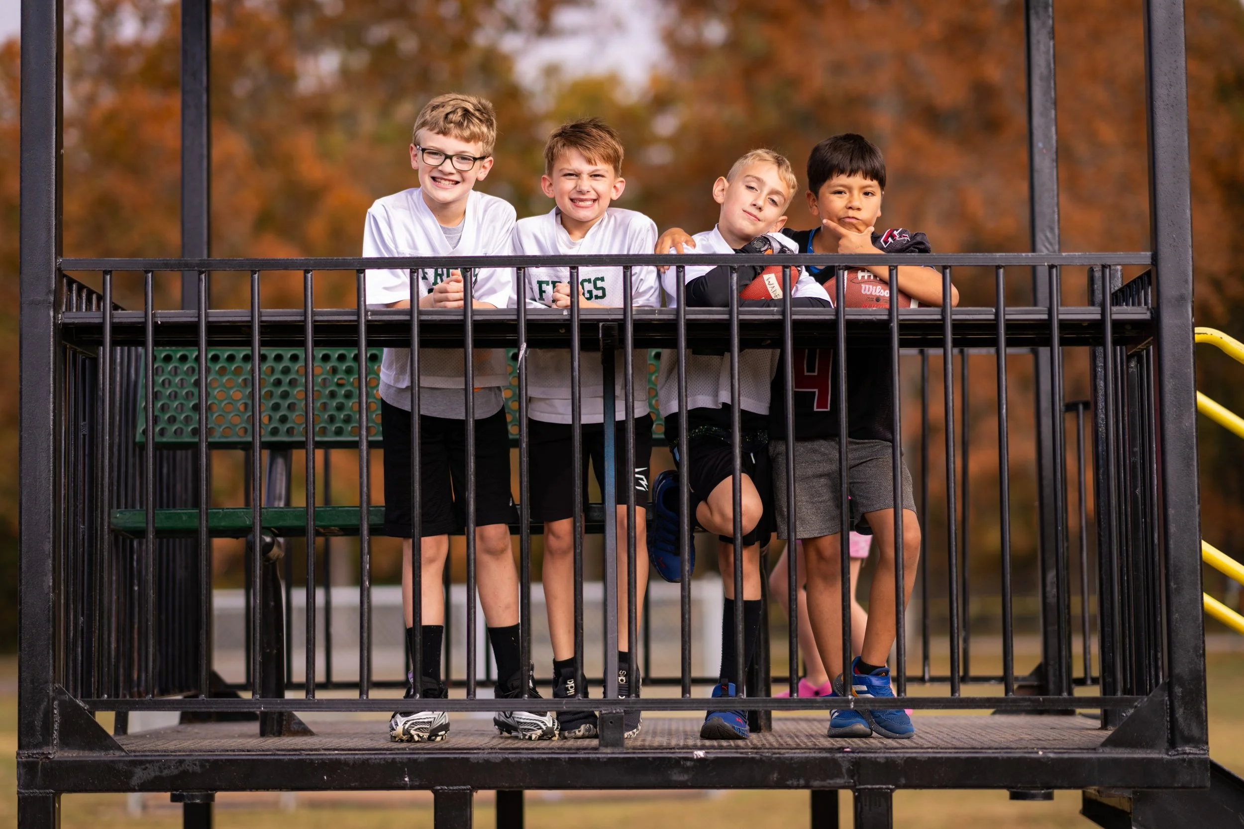 Group of five young boys standing behind a black metal fence at a park, smiling and posing for the camera, with autumn trees in the background.