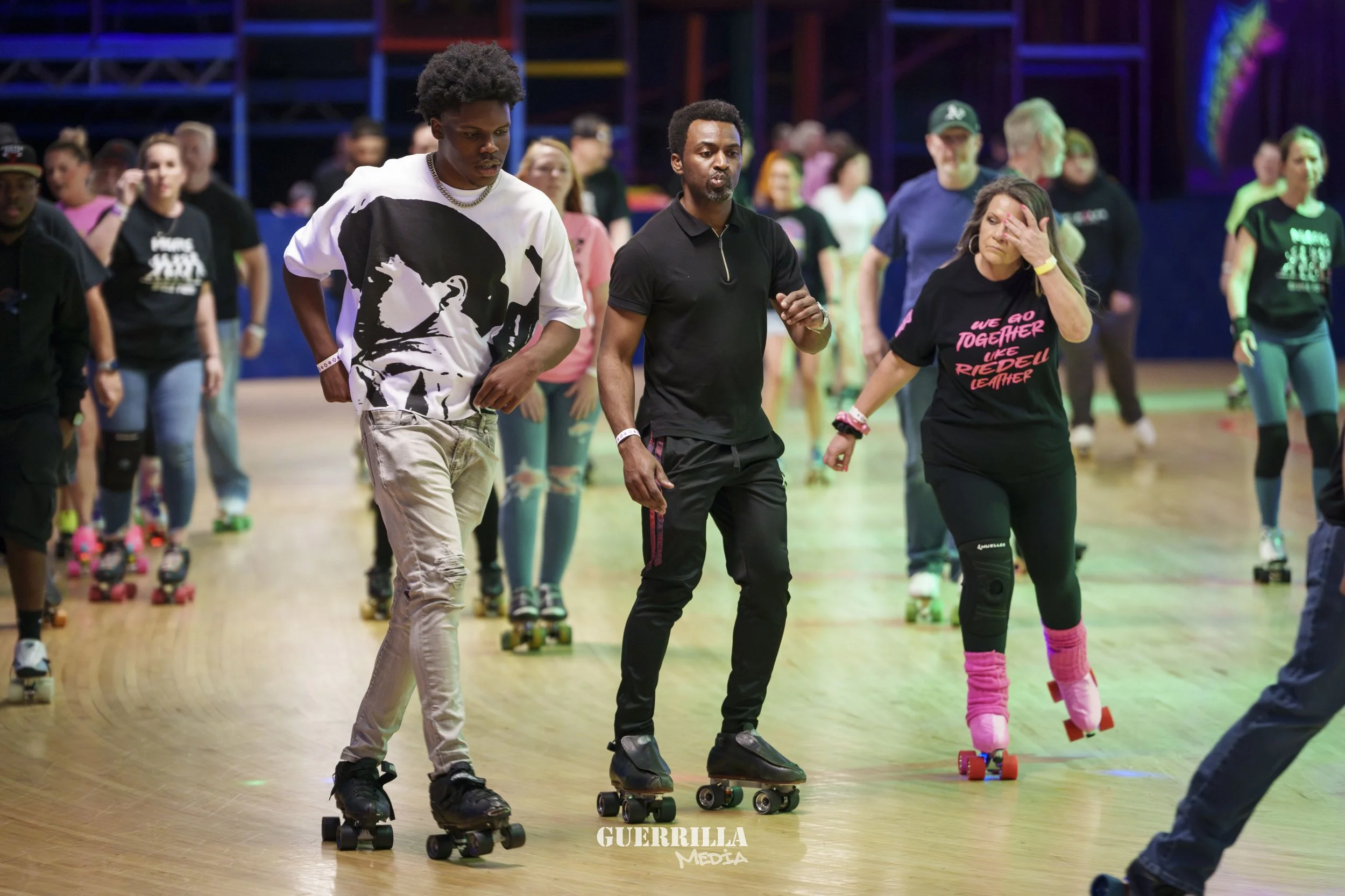 People roller skating indoors, some with protective gear, in colorful lighting with a wooden floor and a crowd in the background.