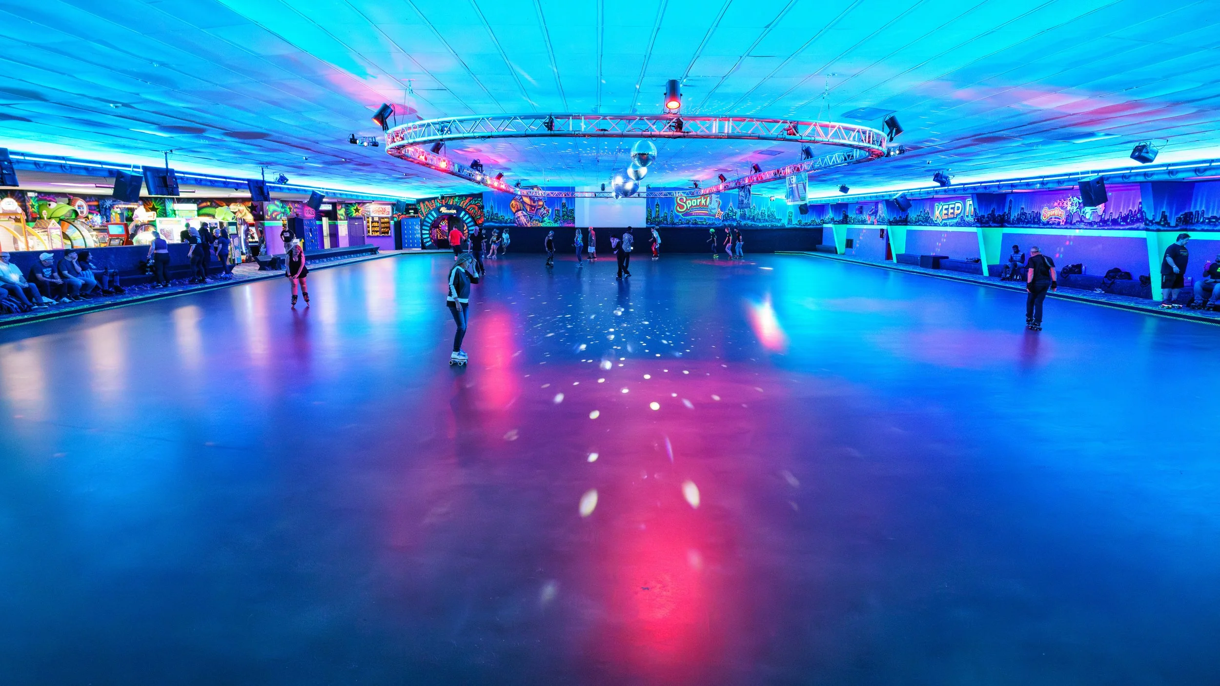 Indoor roller skating rink with colorful blue, pink, and purple lighting, skaters on the rink, and arcade machines along the walls.
