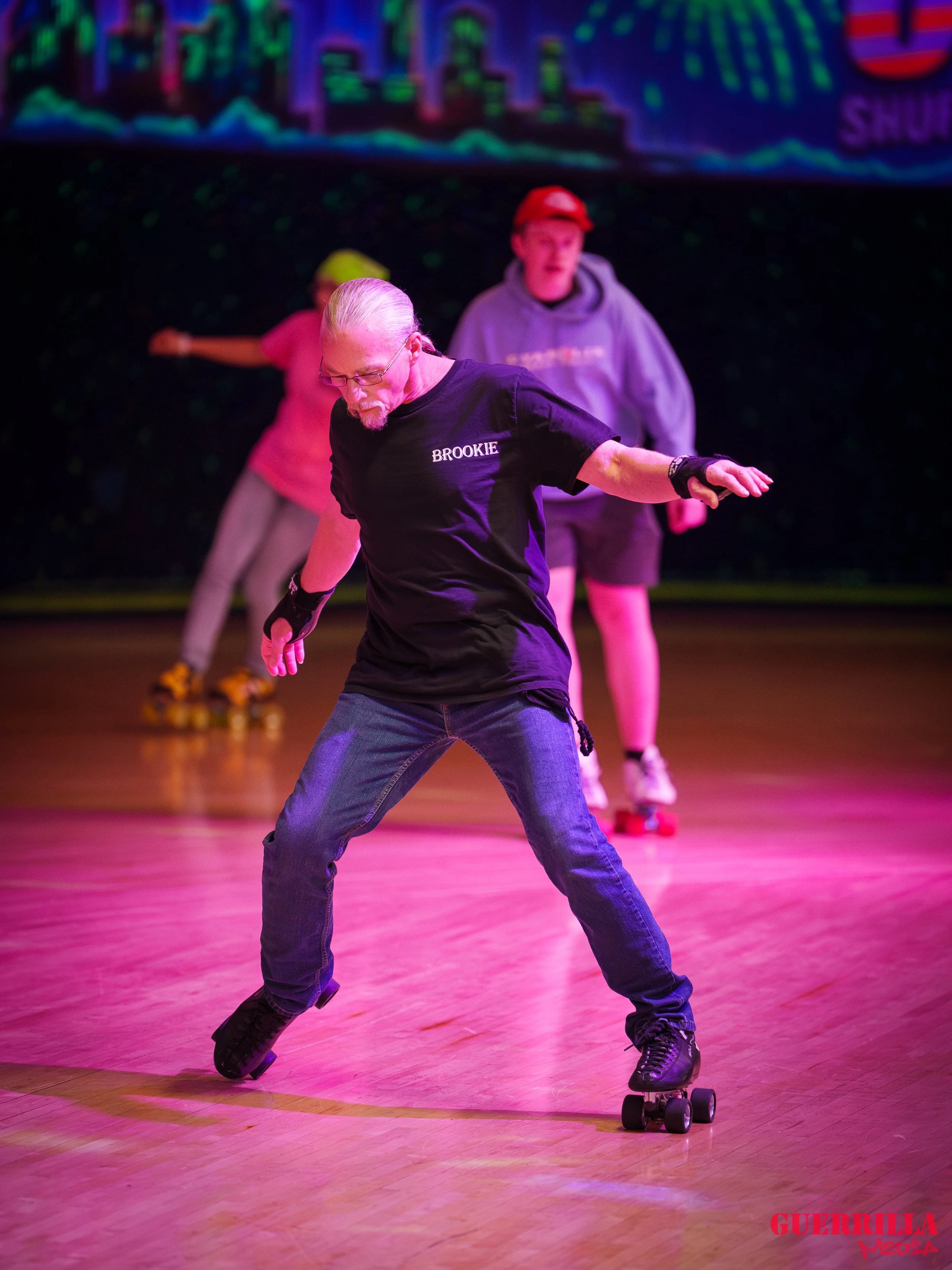 Older man roller skating on a rink with two young people in the background, practicing or performing inline skating.