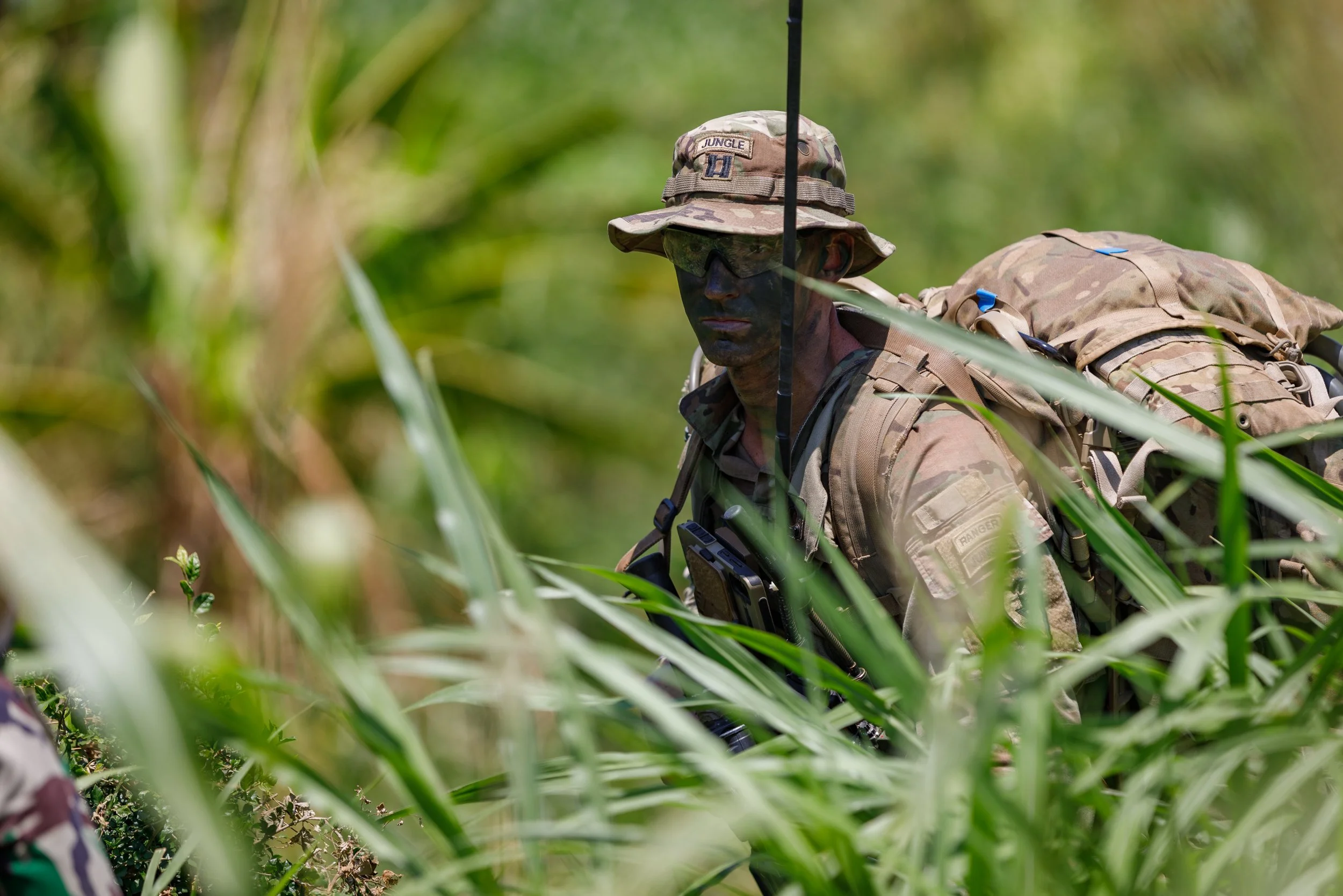 A soldier in camouflage uniform with face paint, wearing a wide-brimmed camouflage hat with a Jungle patch, goggles, and a backpack, navigating through tall grass in a jungle environment.