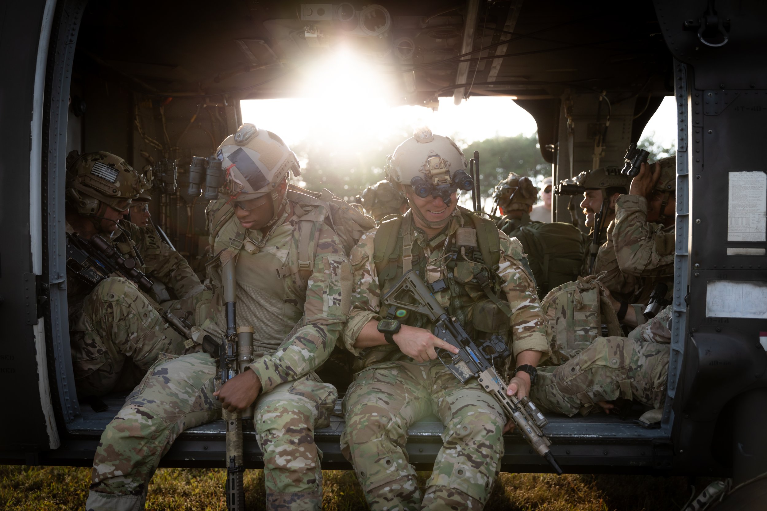 Group of soldiers sitting inside a helicopter, wearing camouflage uniforms and helmets, with sunlight in the background.