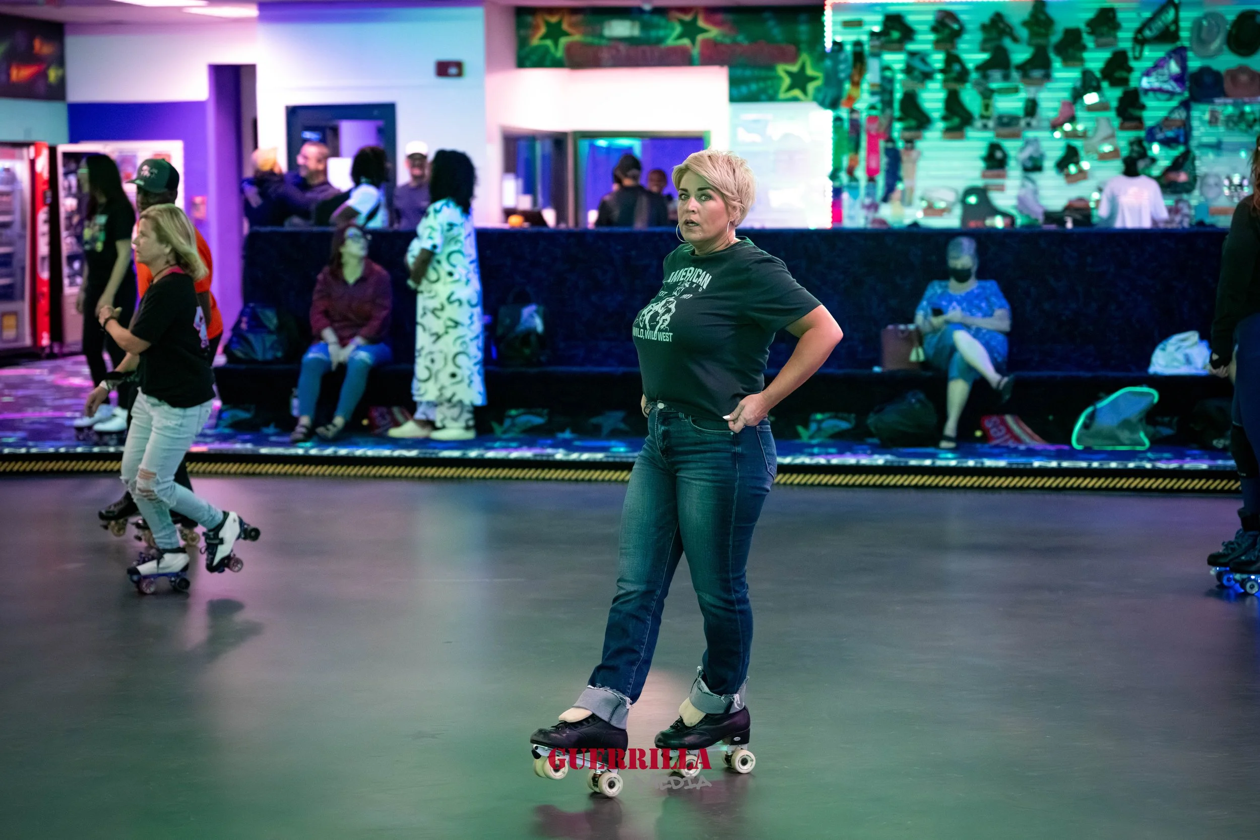 A woman with short blonde hair wearing a black t-shirt and jeans skating in an indoor roller rink with neon lights. Other skaters and people sitting on benches are visible in the background.