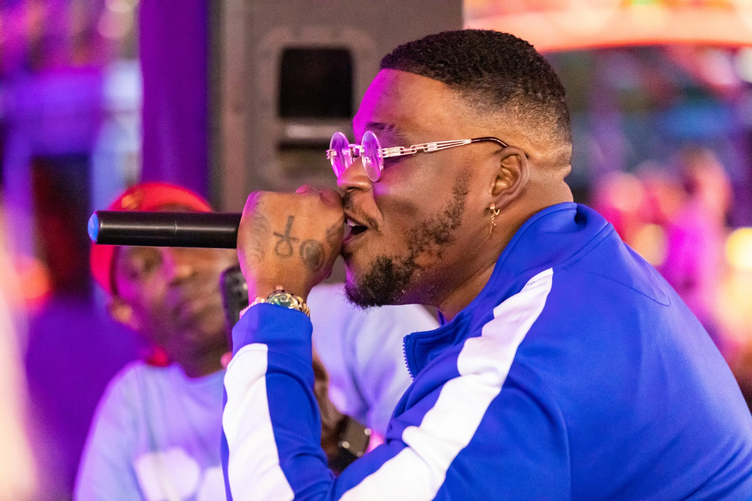 A man wearing a blue and white track jacket is singing into a black microphone at a lively indoor event, with colorful lights and blurred crowd in the background.