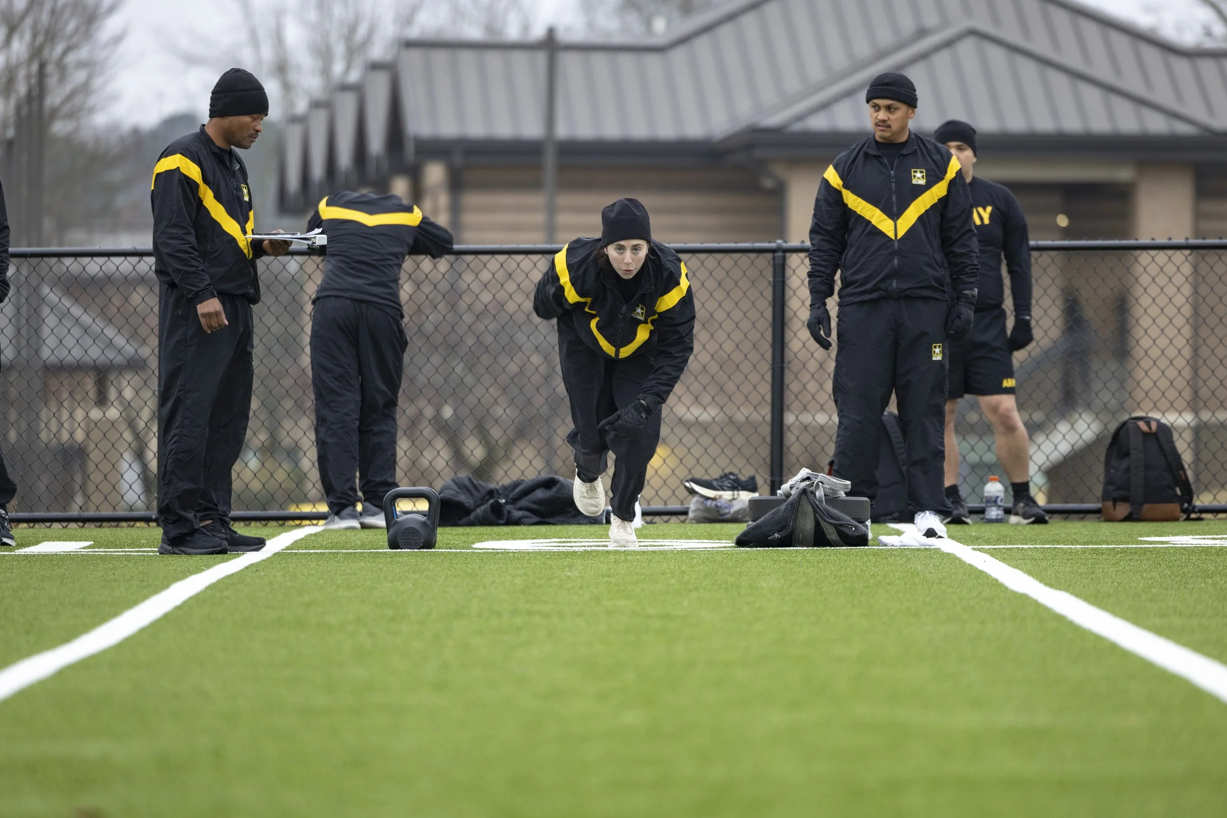 Group of athletes dressed in black and yellow sportswear, preparing on a synthetic field, one athlete is running as part of an exercise, others are standing around with equipment and a coach is writing notes.