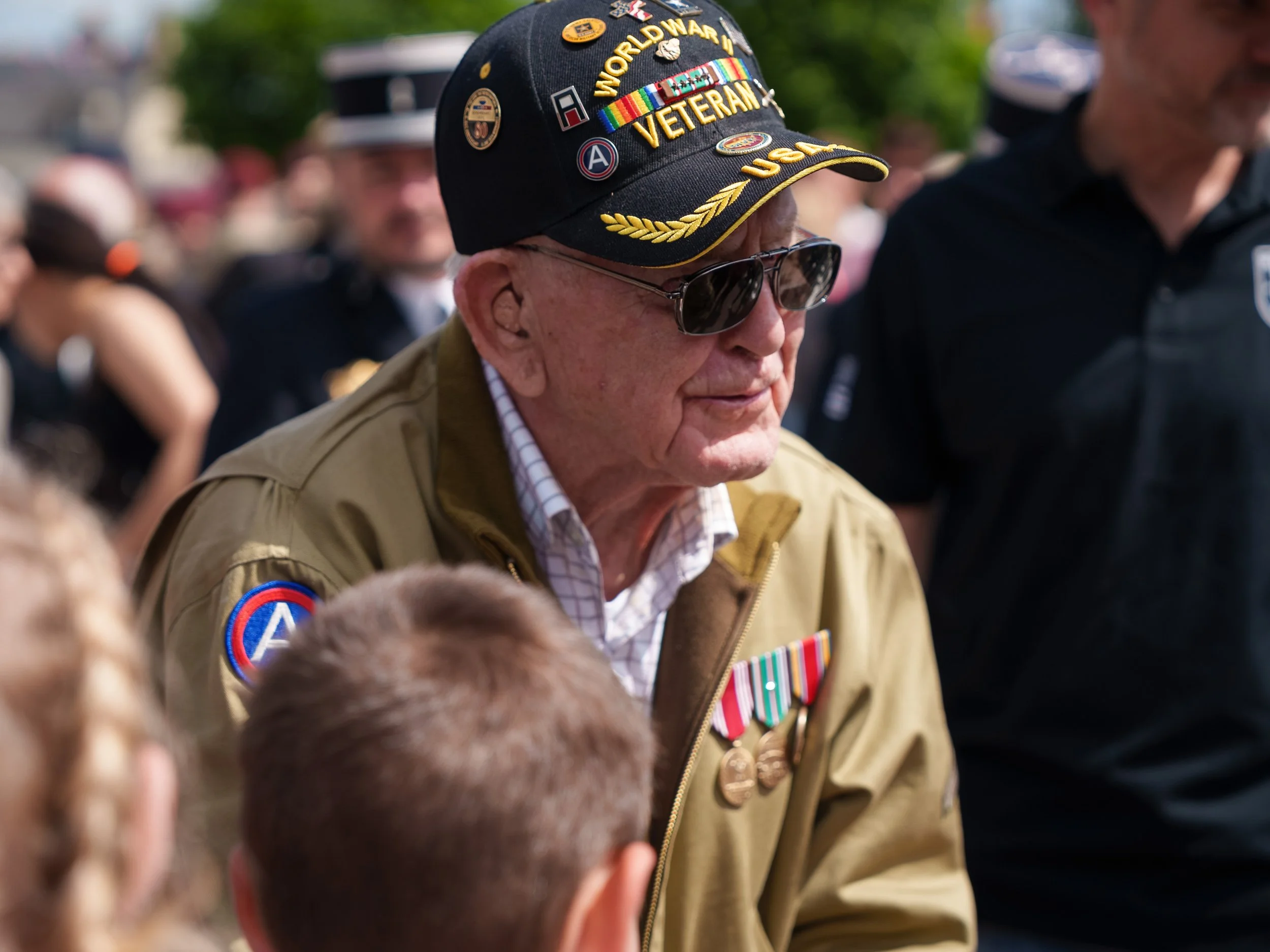 A veteran man wearing a black cap with World War II and U.S. military insignia, sunglasses, a beige jacket with military patches, and medals, at a public event.