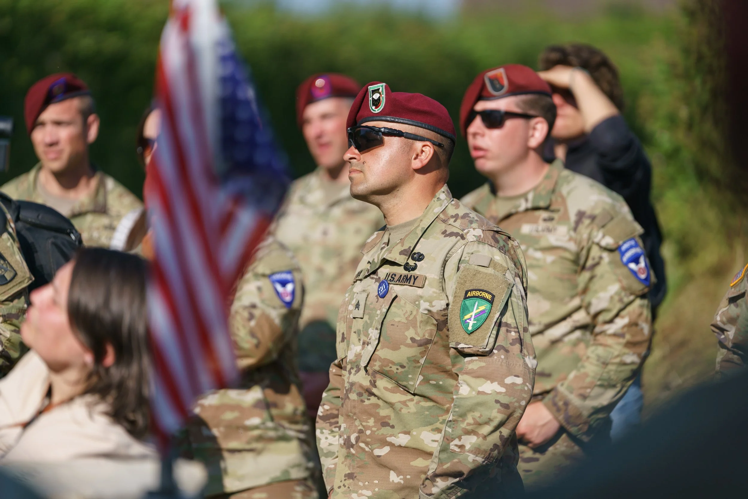 U.S. Army soldiers in camouflage uniforms during a ceremony, with one soldier in a maroon beret and sunglasses in focus, and others in the background, some wearing sunglasses and saluting. An American flag is partially visible in the foreground.