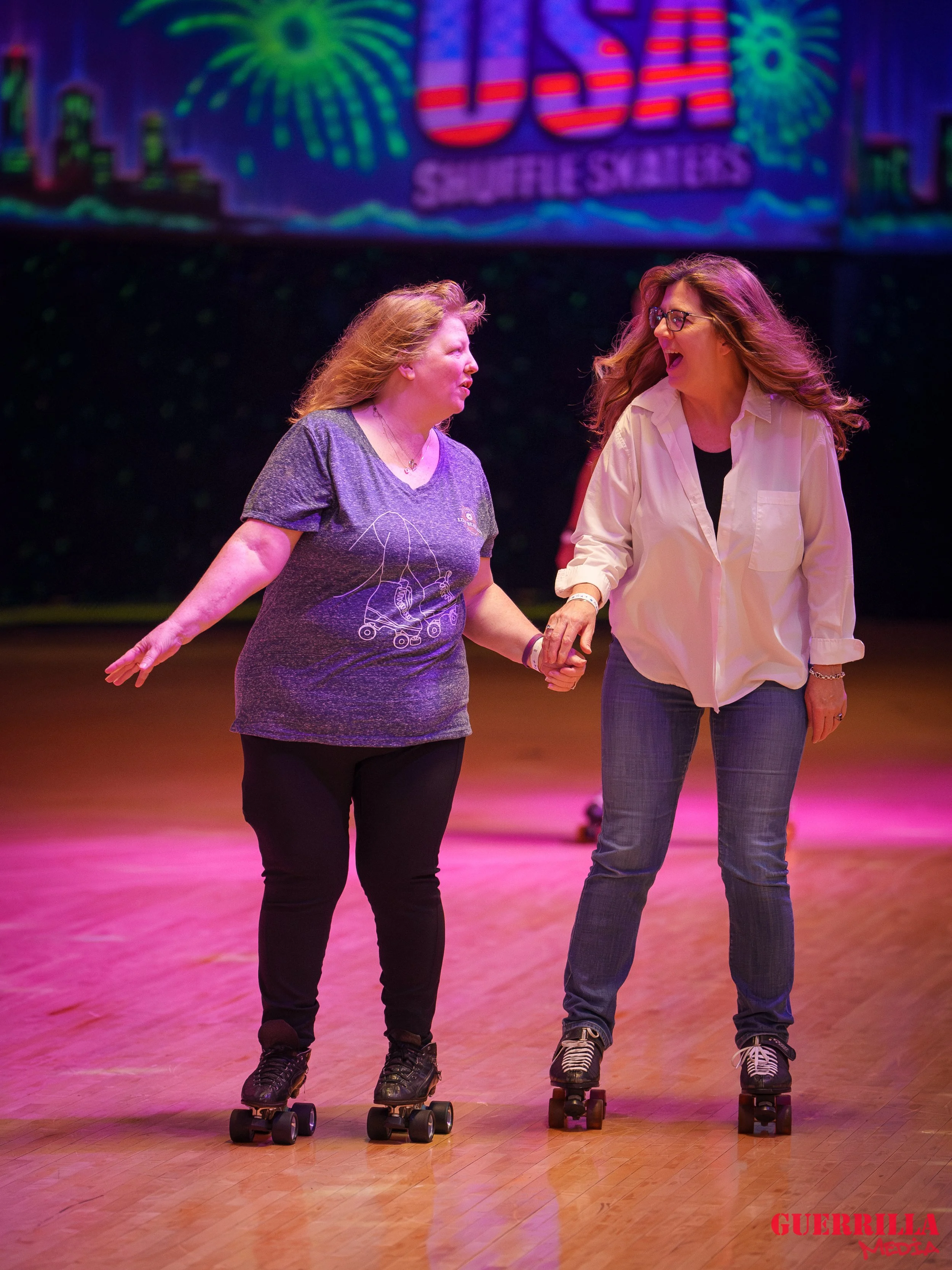 Two women roller skating together on a wooden floor, holding hands, and smiling at each other under colorful stage lights with a performance background.