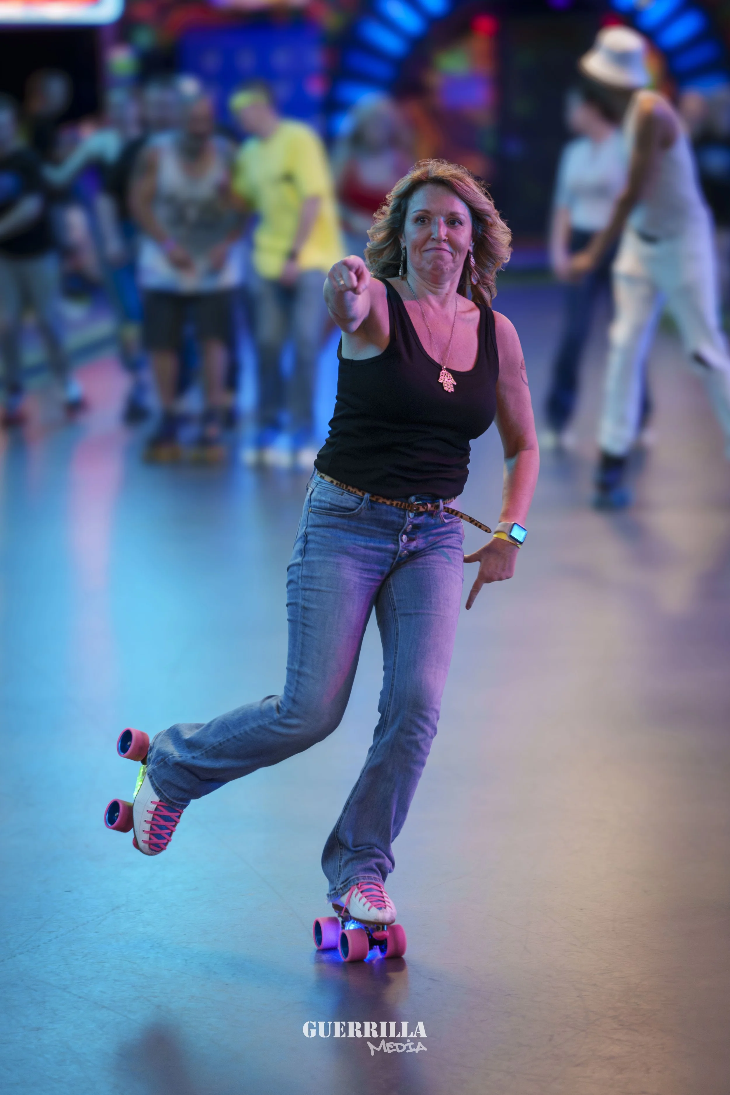 A woman roller skating indoors with a lively background of other skaters and colorful lights, wearing a black tank top, jeans, and a necklace, pointing forward.