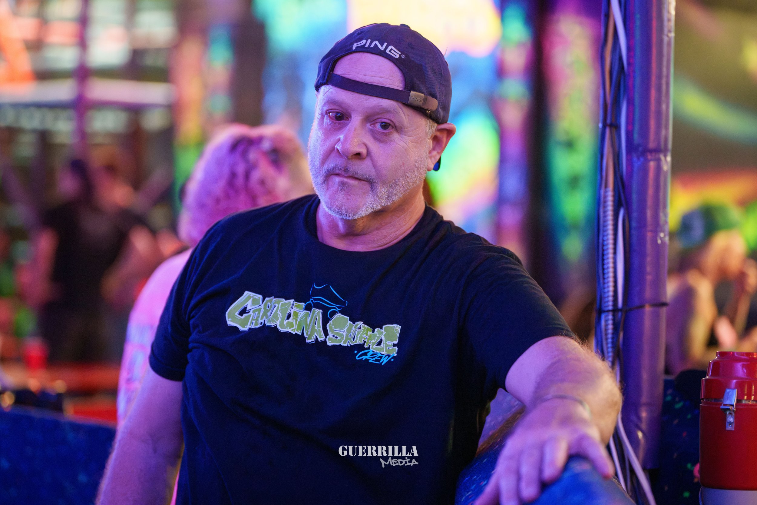 A middle-aged man with a beard and wearing a black 'Green Simple Crew' T-shirt and a backward black cap, standing in a colorful, illuminated environment with blurred people in the background, possibly at a festival or nightclub.