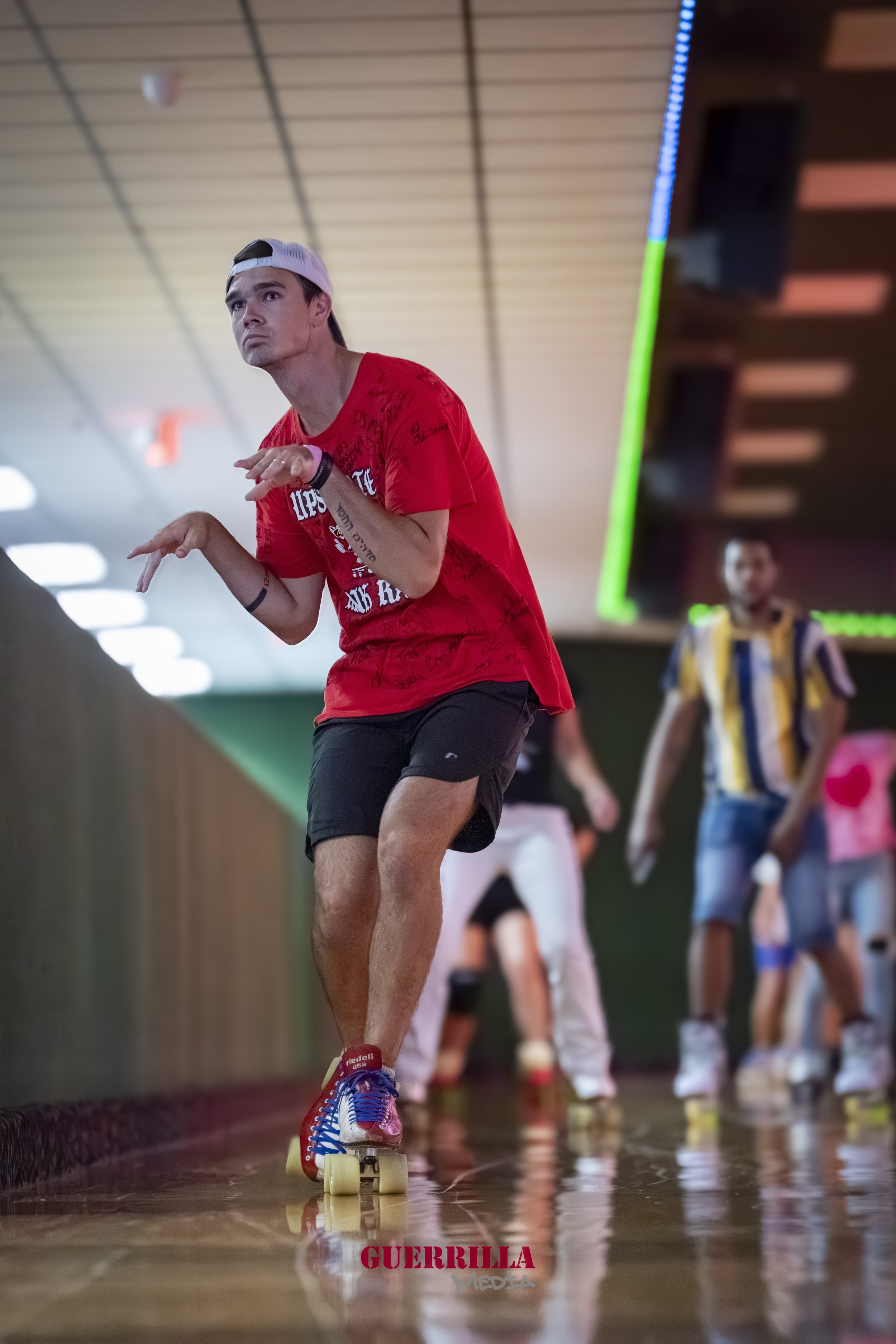 A young man roller skating indoors, wearing a red t-shirt, black shorts, a backward white cap, and a wristband, with others skating behind him in a skating rink.