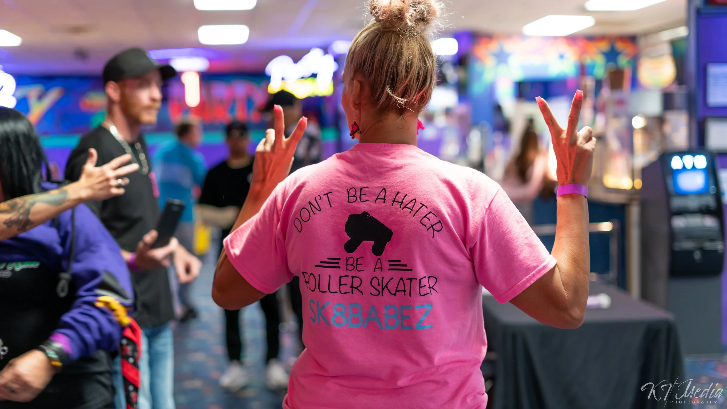 Person with blonde hair in a bun, wearing a pink T-shirt with the message 'Don't Be a Hater, Be a Roller Skater' and peace signs on the back, making peace signs with both hands inside an arcade or entertainment venue.
