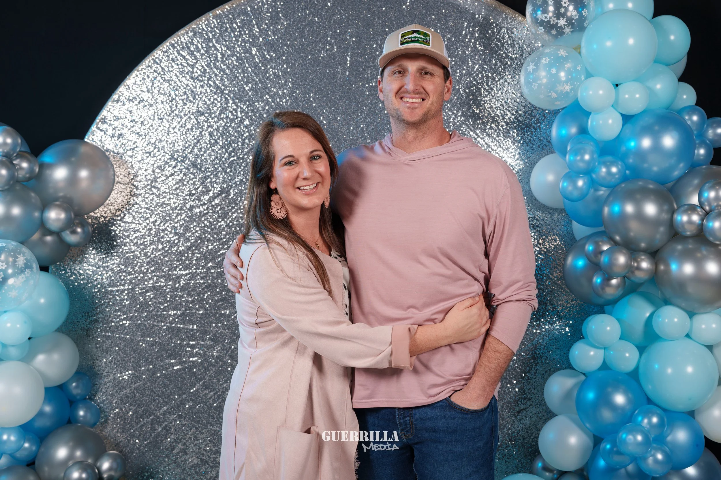 A smiling woman and man standing together in front of a silver glitter backdrop surrounded by blue and silver balloons, with the woman hugging the man.