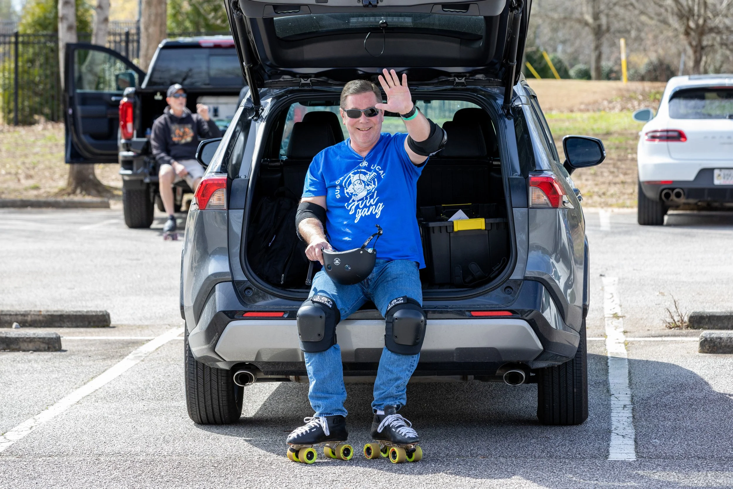 A man sitting on the back of a gray SUV in a parking lot, smiling and waving, wearing roller skates, knee pads, elbow pads, and a blue t-shirt. He is holding a helmet and appears to be enjoying a day of roller skating.