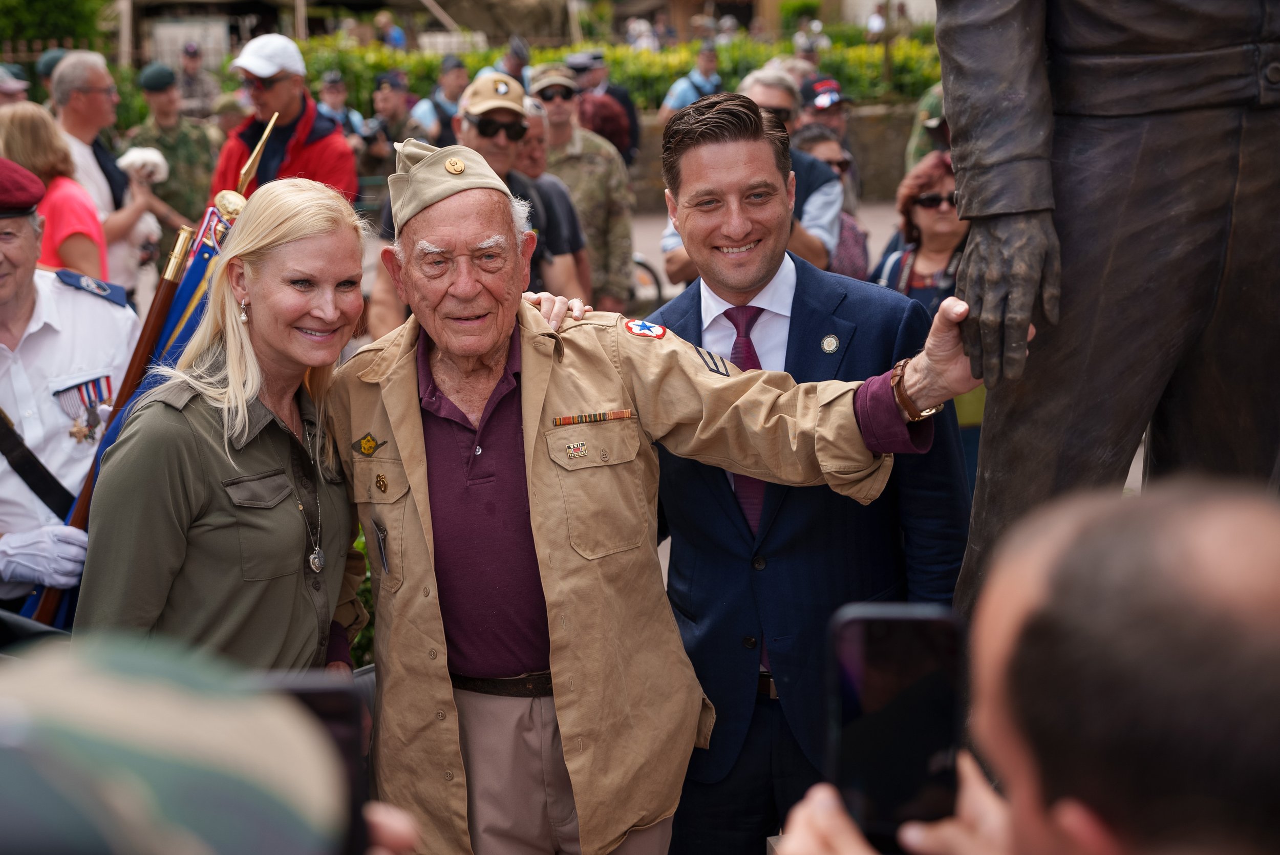A group of people, including a woman and two men, are gathered outdoors, touching a statue's hand during a commemorative event with many spectators in the background.