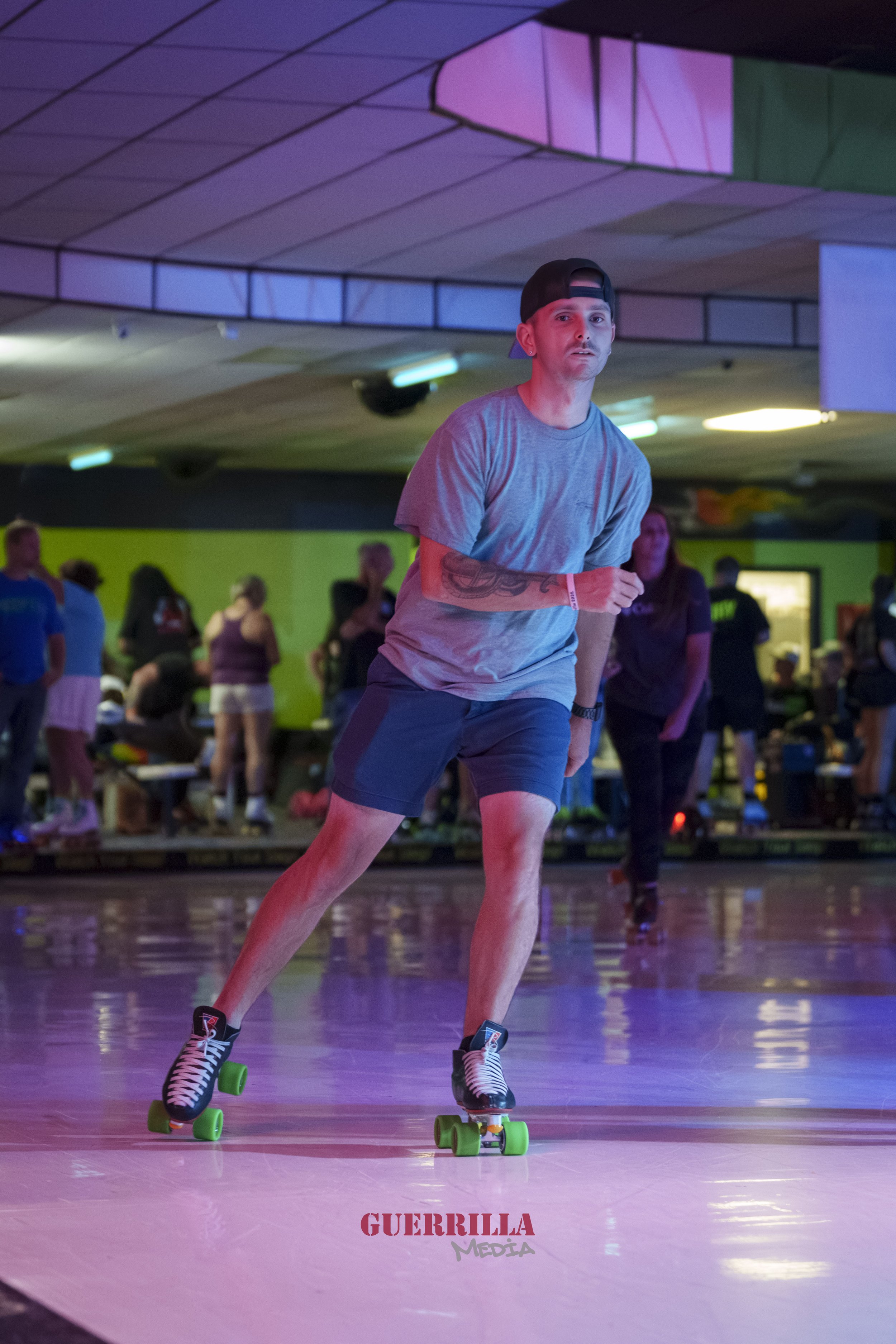 A man roller skating in an indoor roller rink with colorful neon lighting, other skaters and spectators in the background, and a logo that says 'GUERRILLA MEDIA' on the floor.