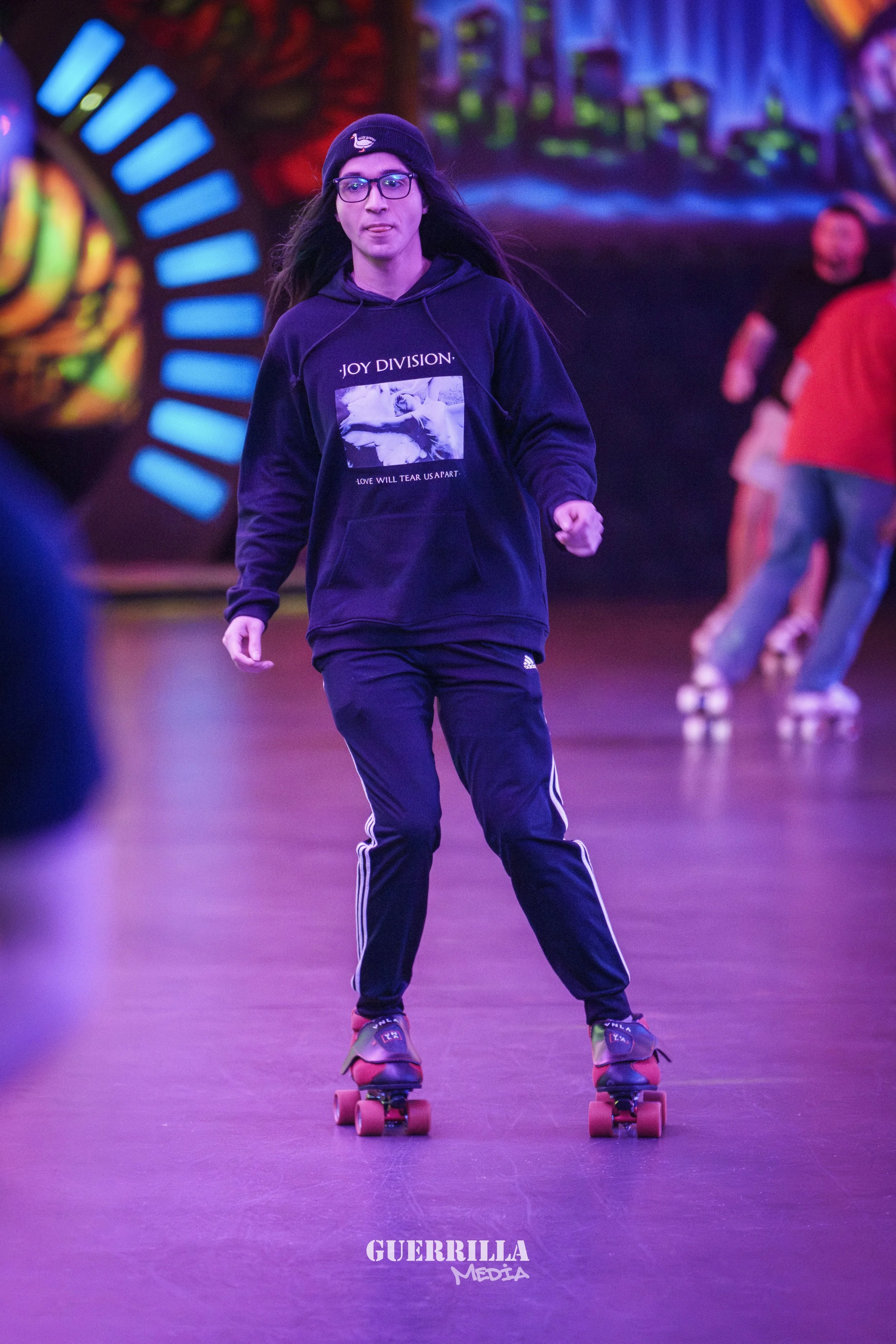 A person roller skating indoors, wearing glasses, a beanie hat, a black hoodie with 'Joy Division' on it, and black Adidas pants, with a colorful, vibrant background.