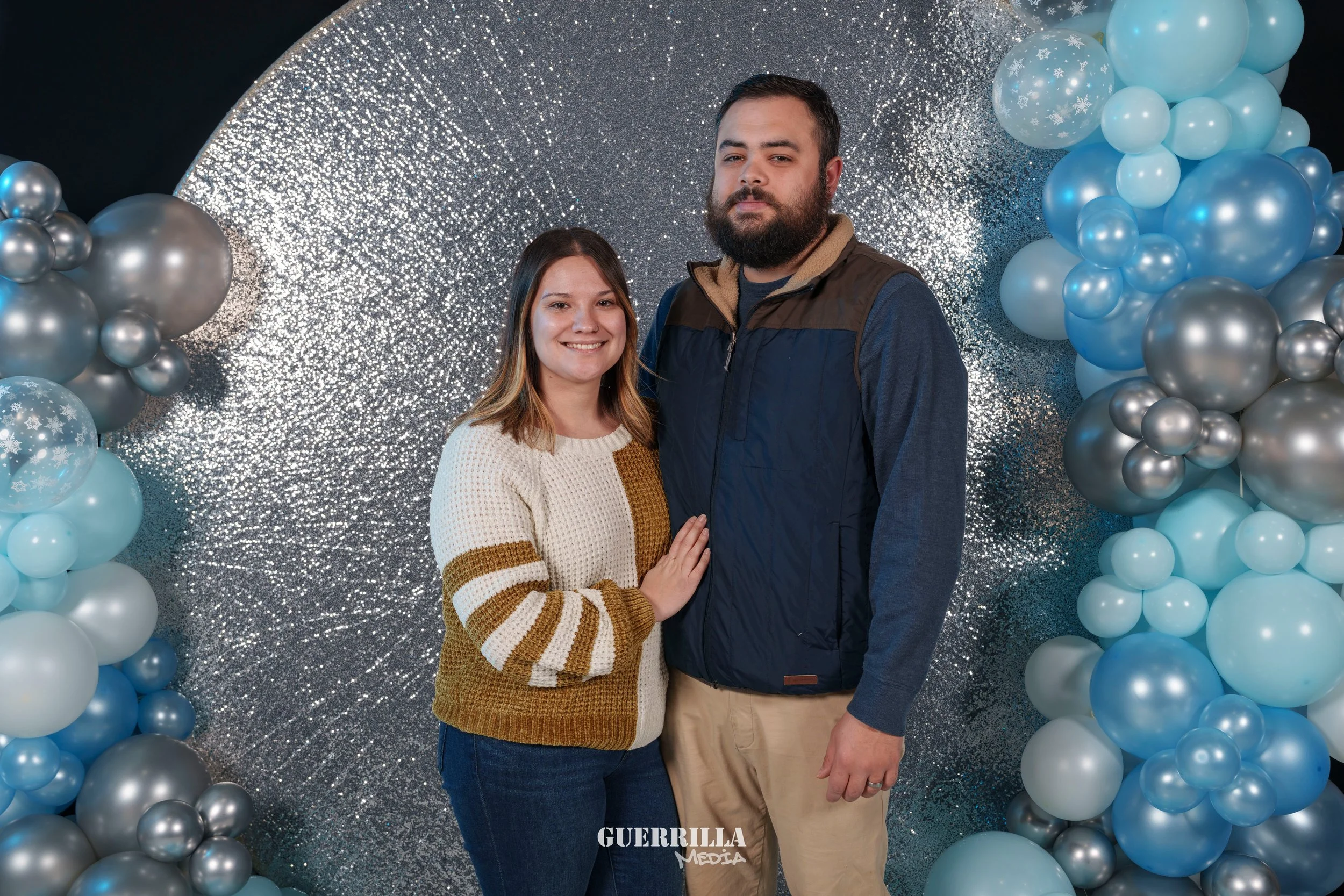 A smiling couple standing in front of a festive backdrop with balloons and sparkly background, posing together.