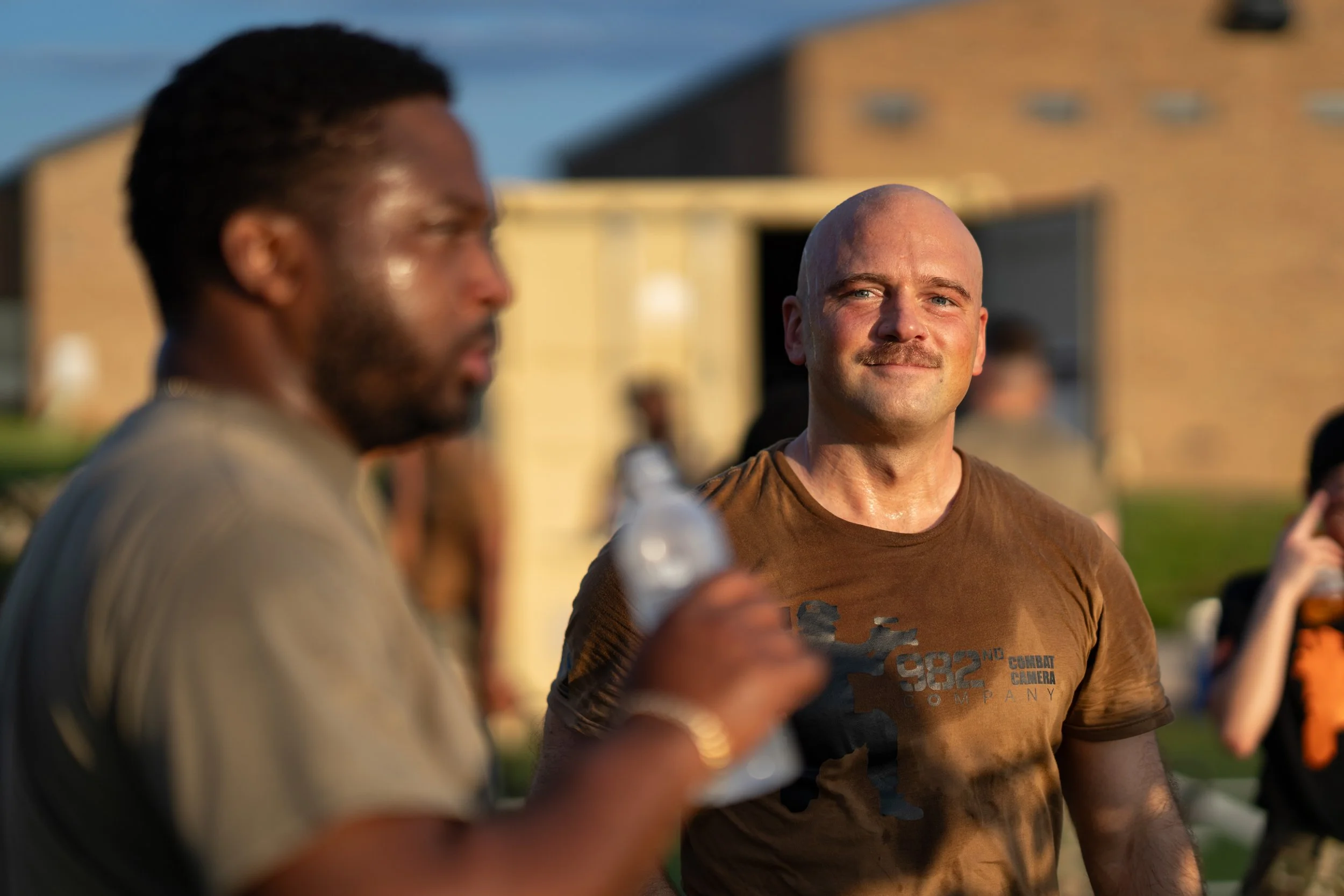 Two men standing outdoors during the day, one is looking forward with a slight smile, the other is blurred in the foreground holding a water bottle, both appear sweaty, with a group of people and a building in the background.