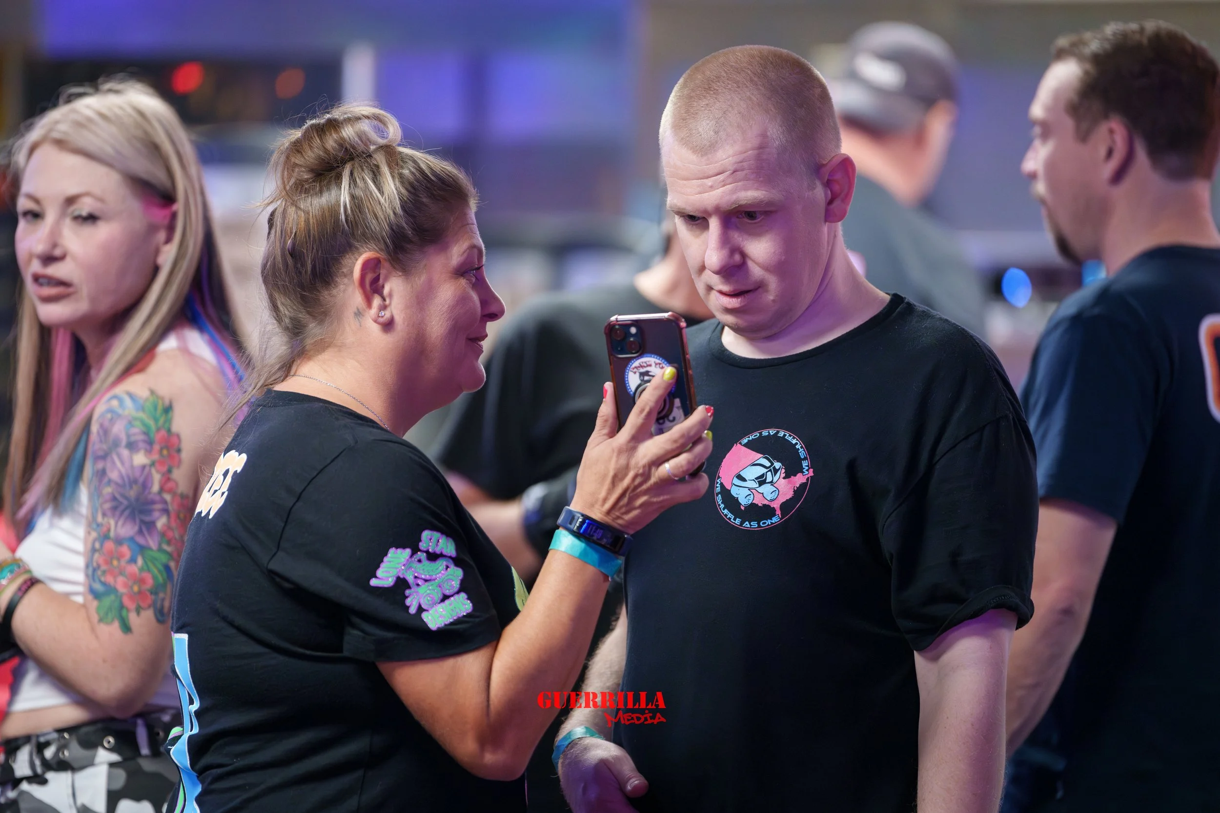 A group of people at a social gathering, with two women and two men in focus. The woman on the left has tattoos and colorful hair streaks, the woman in the center is showing something on her phone to the man with a buzz cut, who is looking at the pho