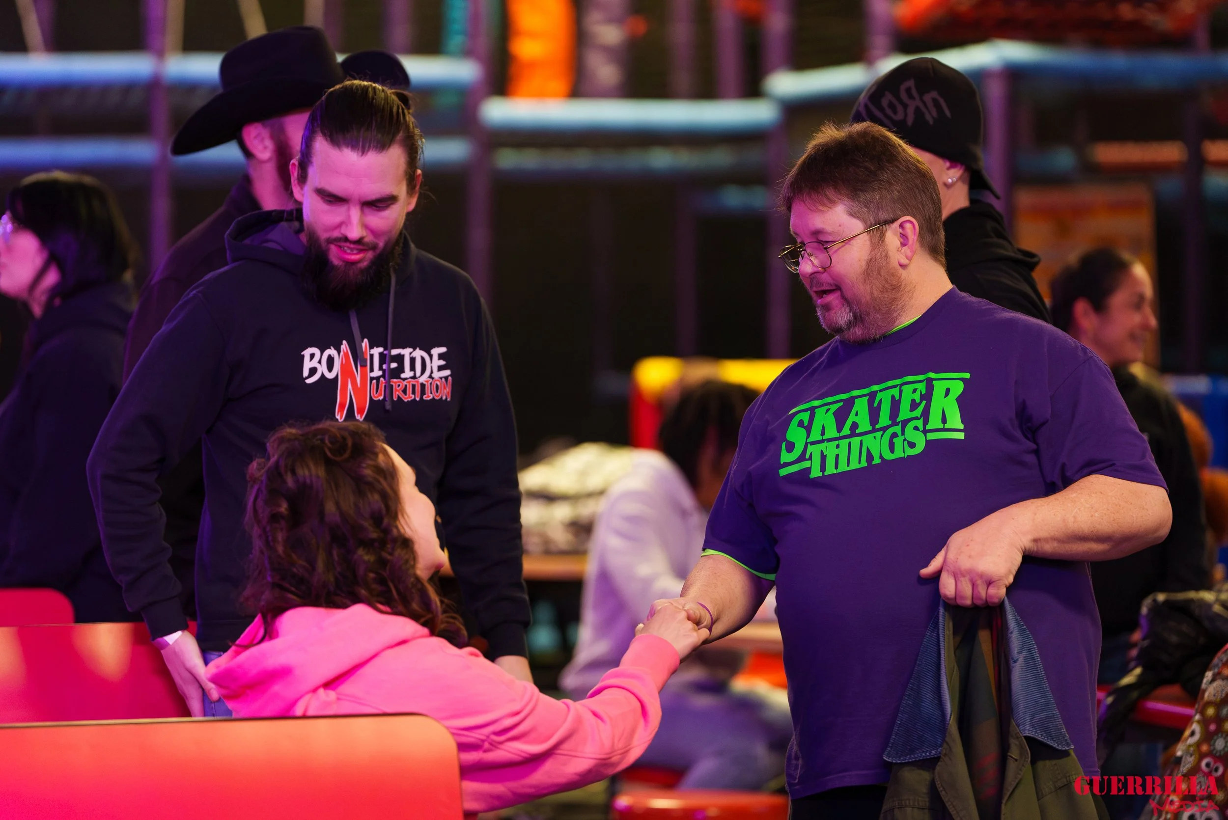Two men and a woman engaging in a handshake and conversation at a social event. The woman is seated wearing a pink hoodie, and the man standing is wearing a blue t-shirt with green text that says "Skater Things." The other man is wearing a black hood