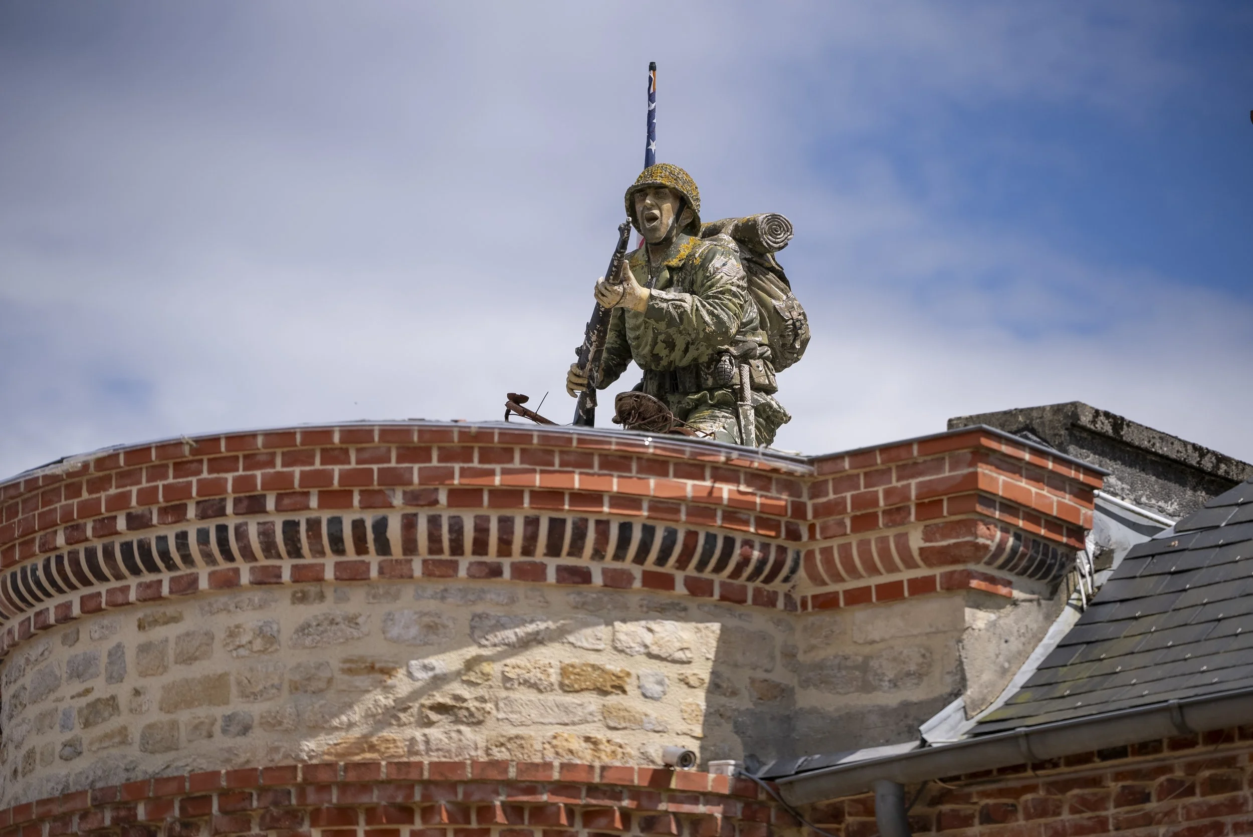 A statue of a soldier in camouflage uniform with a backpack and rolled-up mat, holding a rifle, positioned on top of a brick building against a cloudy sky.