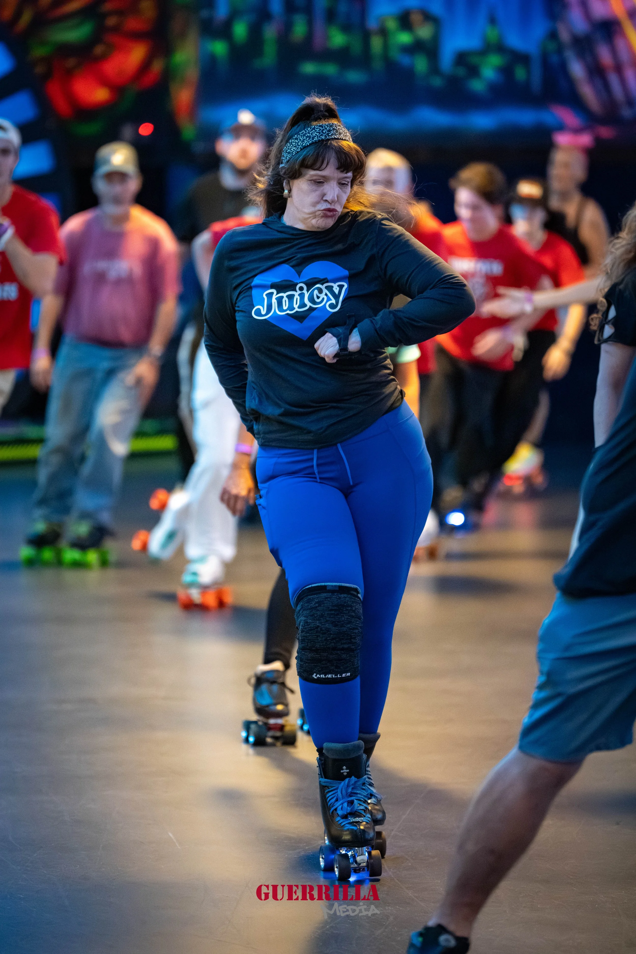 A woman roller skating indoors, wearing a black Juicy hoodie, blue pants, and a knee brace, with a group of people in the background.