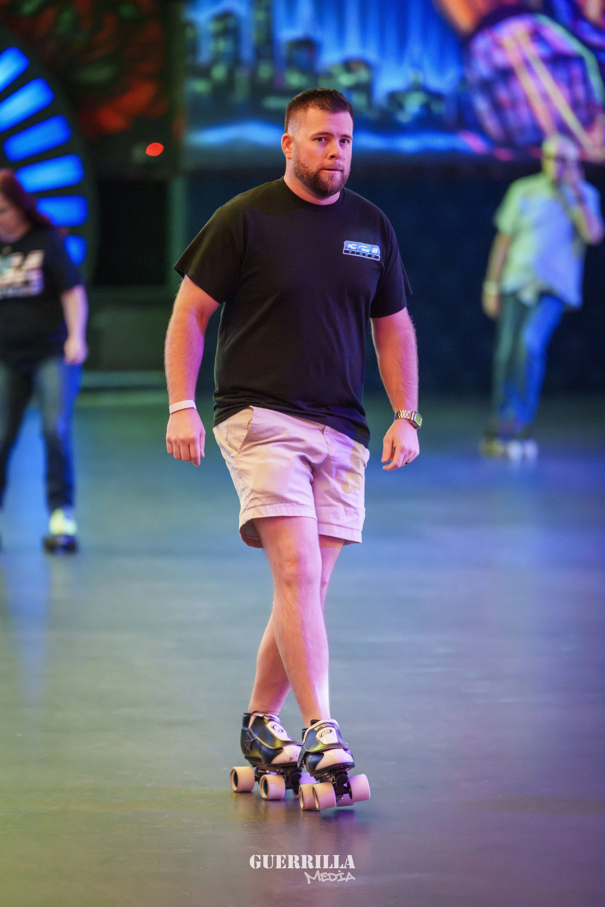 A man roller skating indoors at an amusement center, wearing a black T-shirt, beige shorts, and black roller skates. There are colorful lights and blurred people in the background.