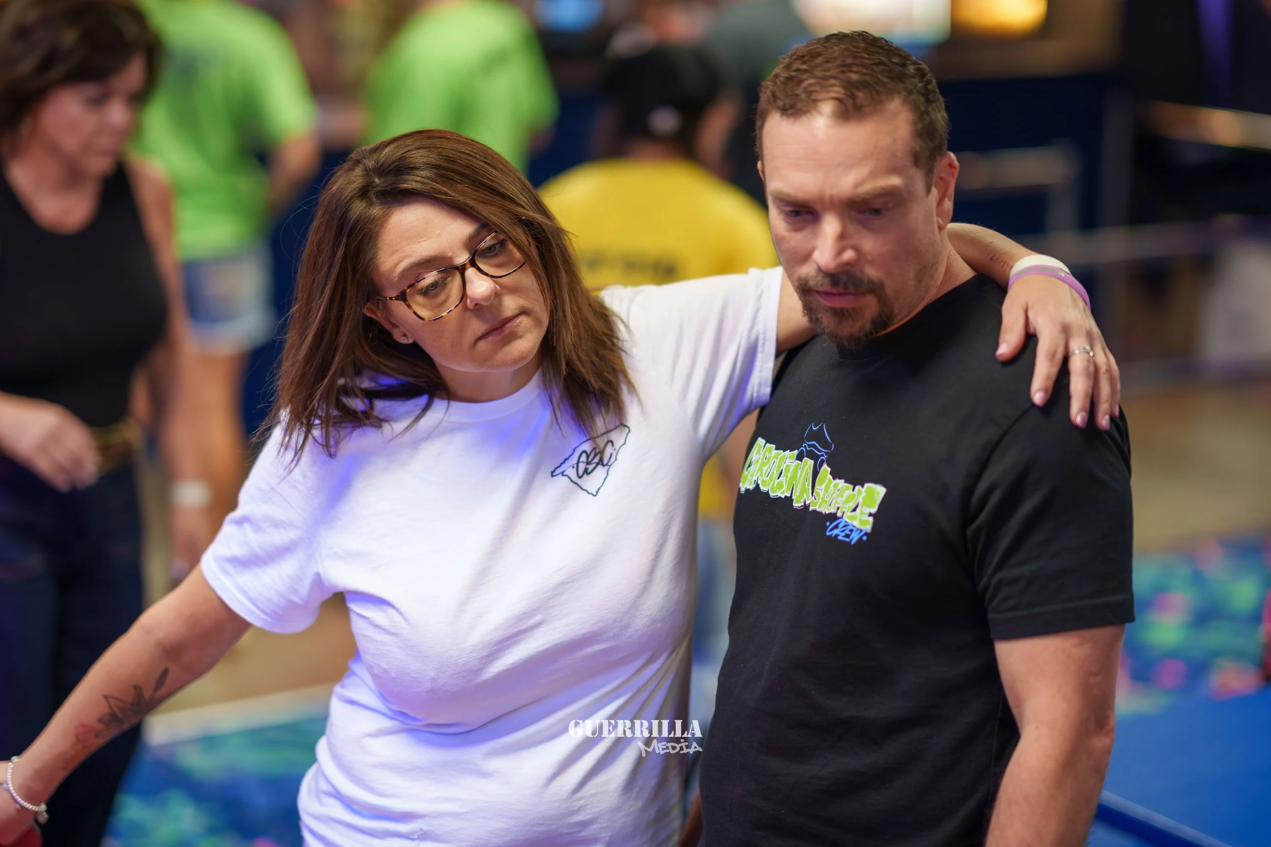 A woman with glasses and a tattoo on her arm helping a man who appears to be injured, at an indoor event with other people in the background.