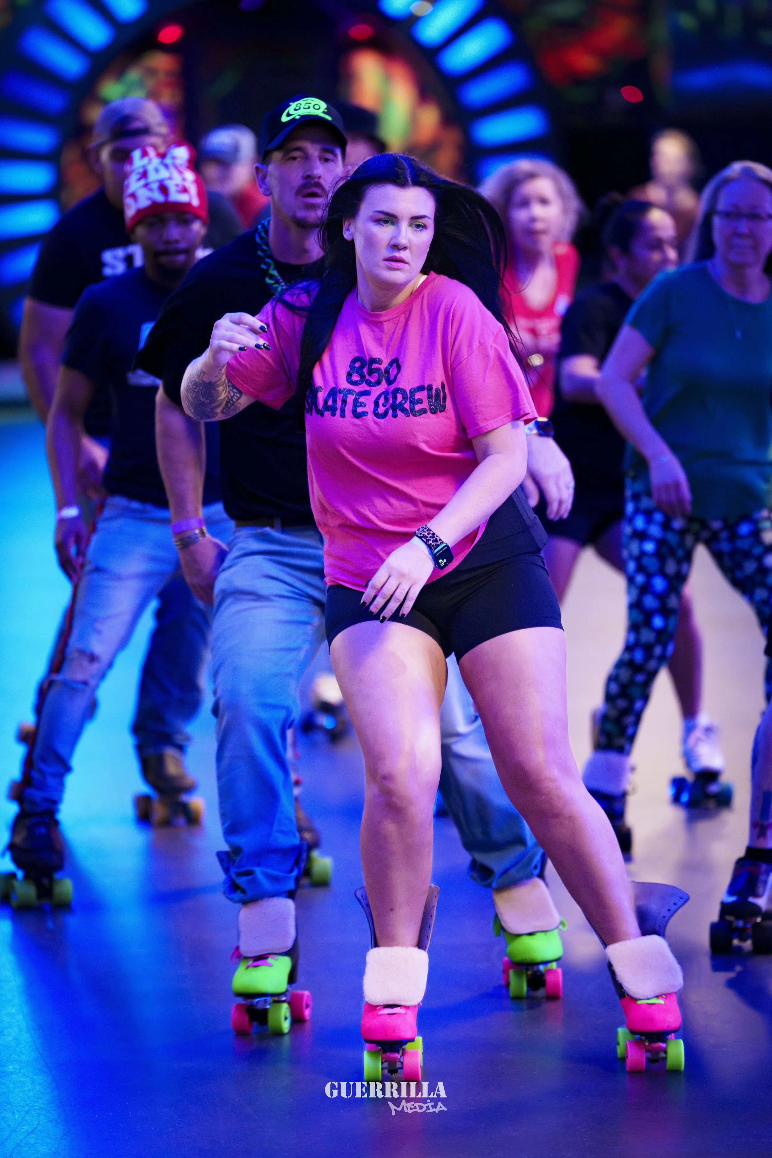 A group of people roller skating indoors, including a woman in a pink shirt that says '850 SKATE CREW,' in the foreground, with others following behind, all skating on a dark floor with colorful lighting.