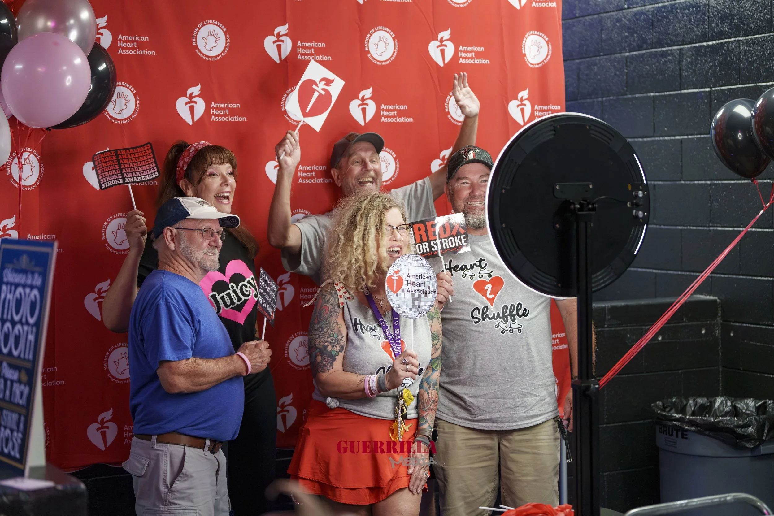 Group of people participating in a Heart Association event, smiling and holding signs supporting stroke awareness, in front of a red backdrop with the American Heart Association logo, some wearing themed shirts and accessories.